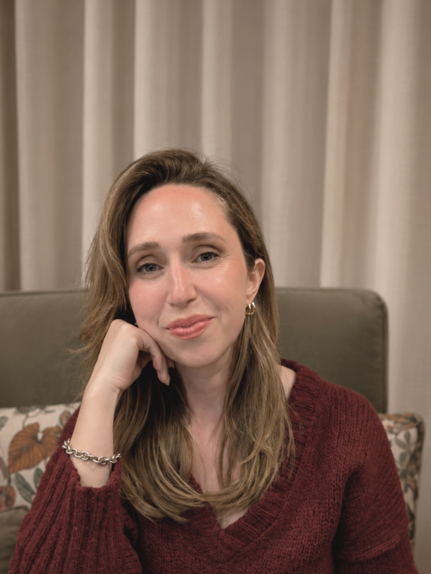 A woman with shoulder-length brown hair wearing a maroon sweater, gold earrings, and a silver bracelet, sitting on a sofa with beige curtains in the background.