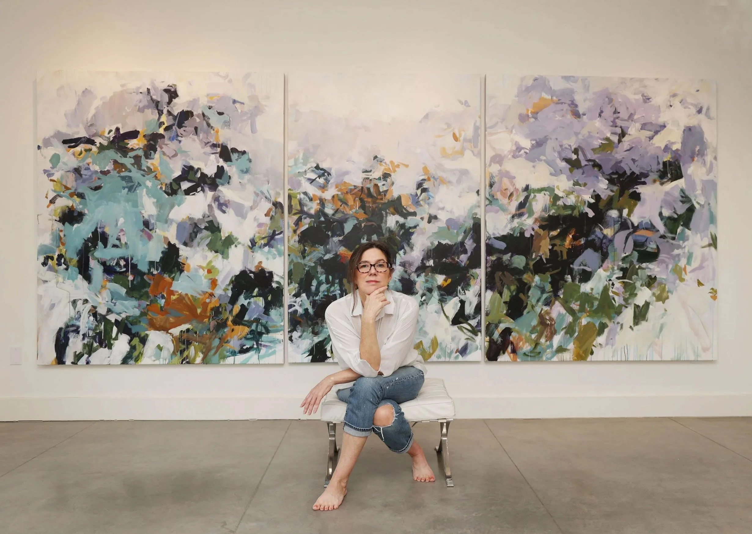 Artist Karen Silve sitting on a white bench in front of one of her large abstract paintings in triptych, with her chin resting on her hand and her left leg crossed over her right knee, dressed in a white shirt and ripped jeans.