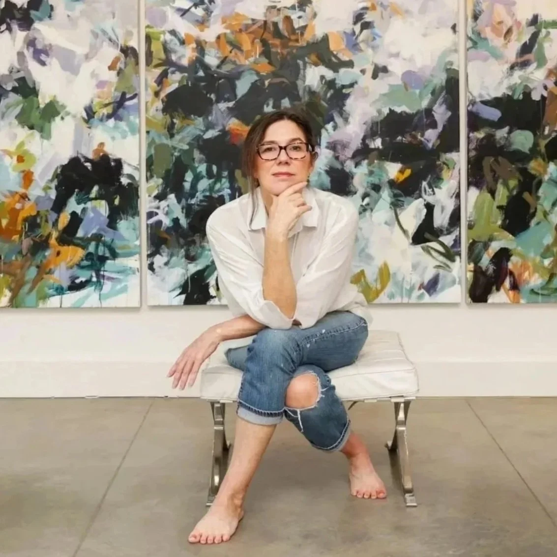 Artist Karen Silve sitting on a white bench in front of three of her abstract paintings, wearing glasses, a white shirt, and ripped jeans, with her leg crossed and her hand on her chin.