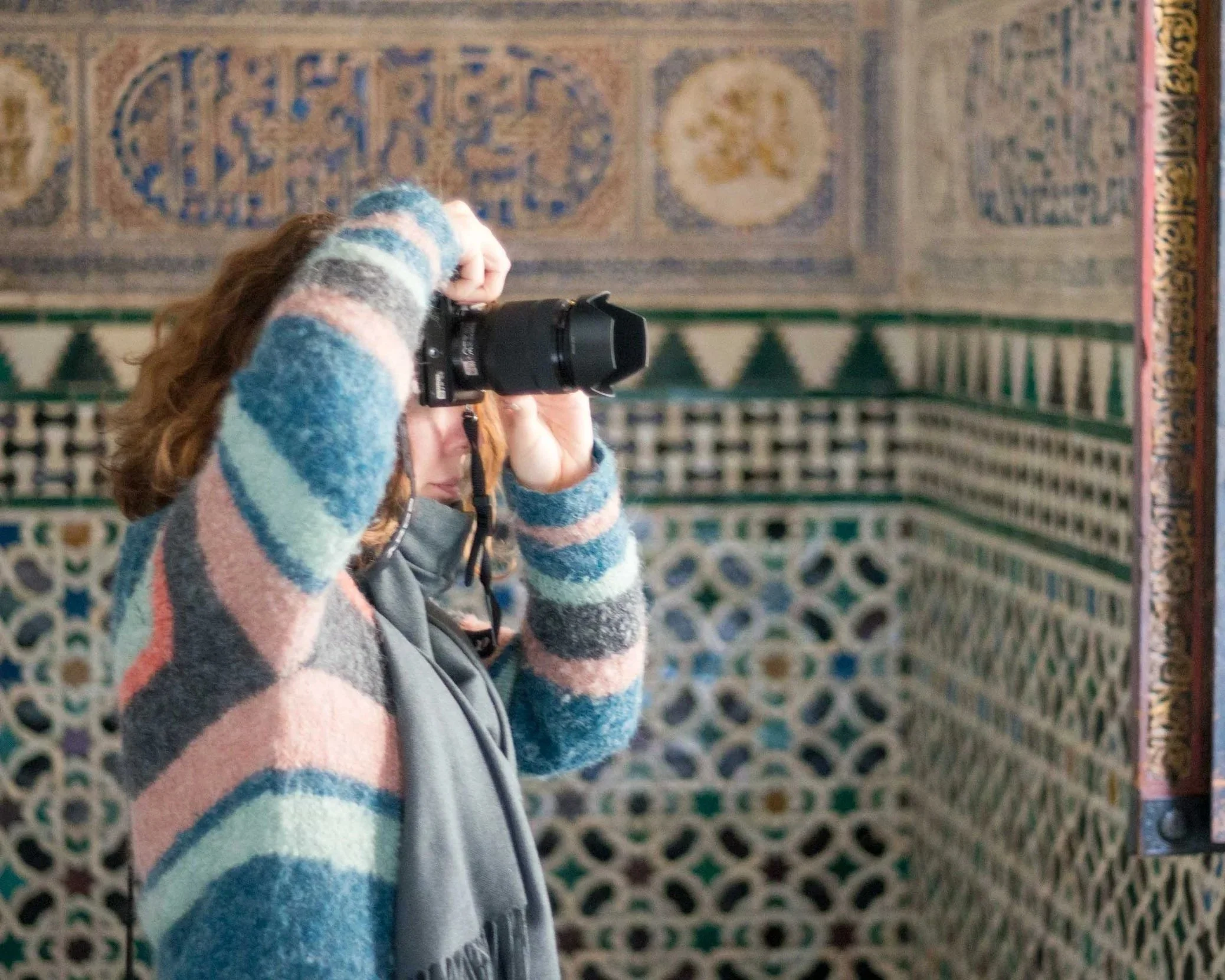 Artist and photographer Kimberly Poppe at work, holding a camera, wearing a colorful striped sweater, standing in front of a patterned tile wall.