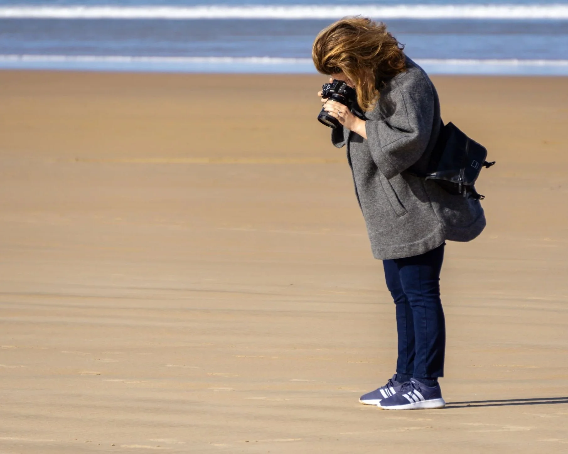 Kimberly Poppe, fine art photographer, on the beach taking a photograph with a camera, wearing a gray coat, dark jeans, and sneakers, with ocean waves in the background.