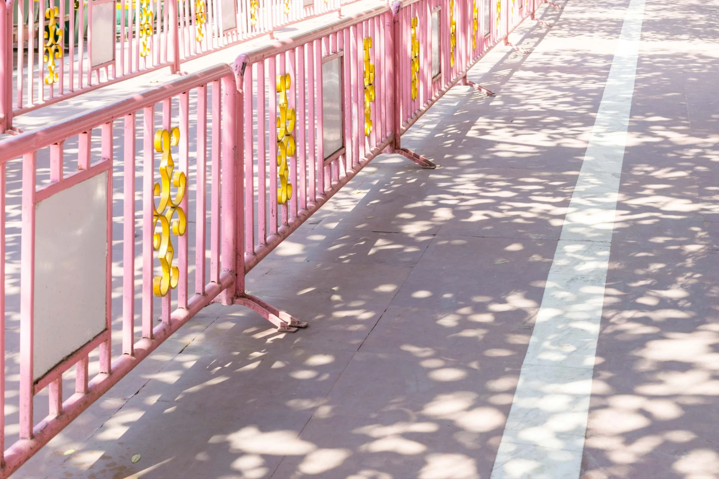 Contemplative (Miksang) photograph by artist Kimberly Poppe. Pink metal barricades with yellow decorative elements cast shadows on the sidewalk near a white line. The scene is outdoors, illuminated by sunlight.