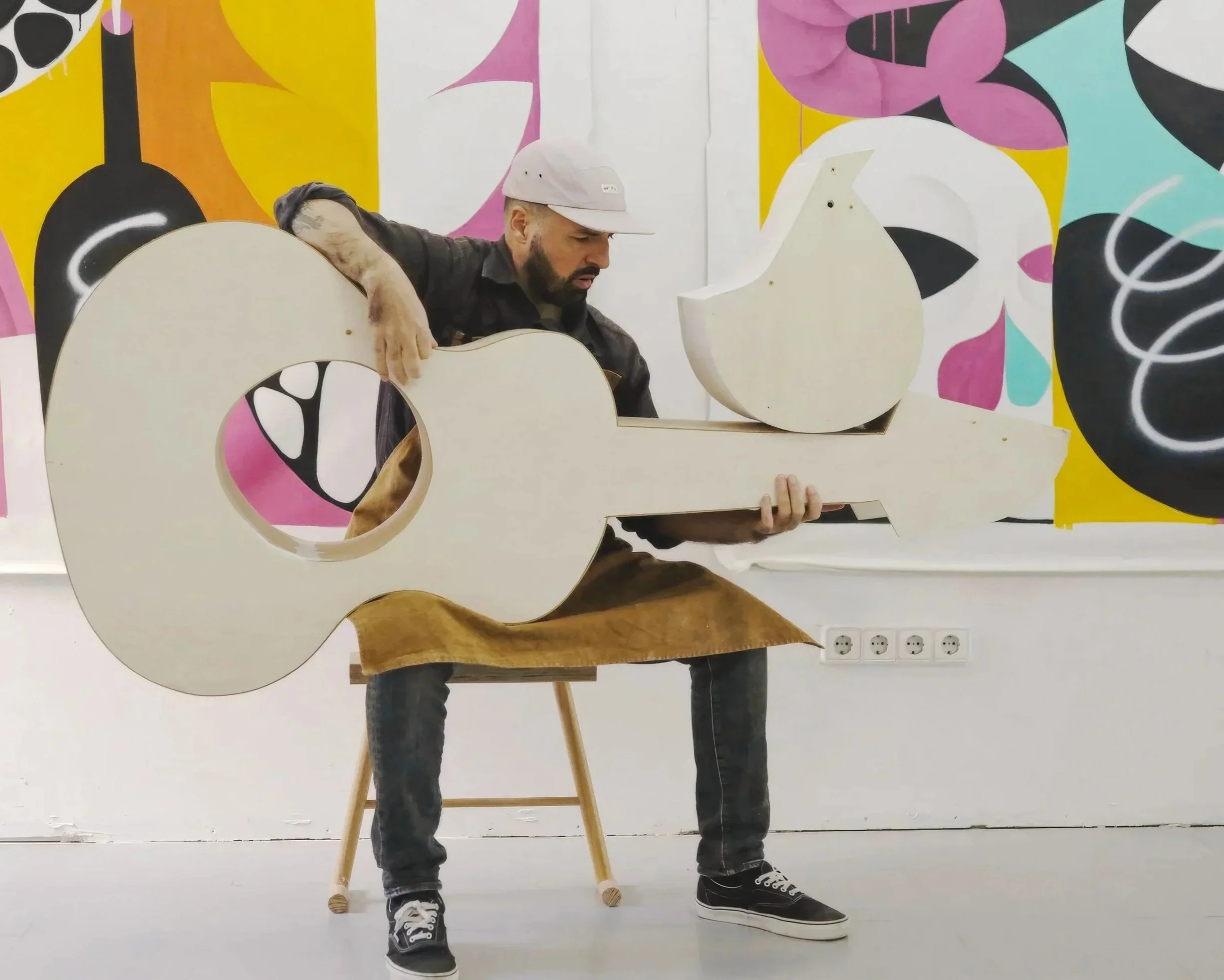 Artist Ruben Sanchez assembling a large abstract wooden sculpture with geometric cutouts, set against a colorful mural background.