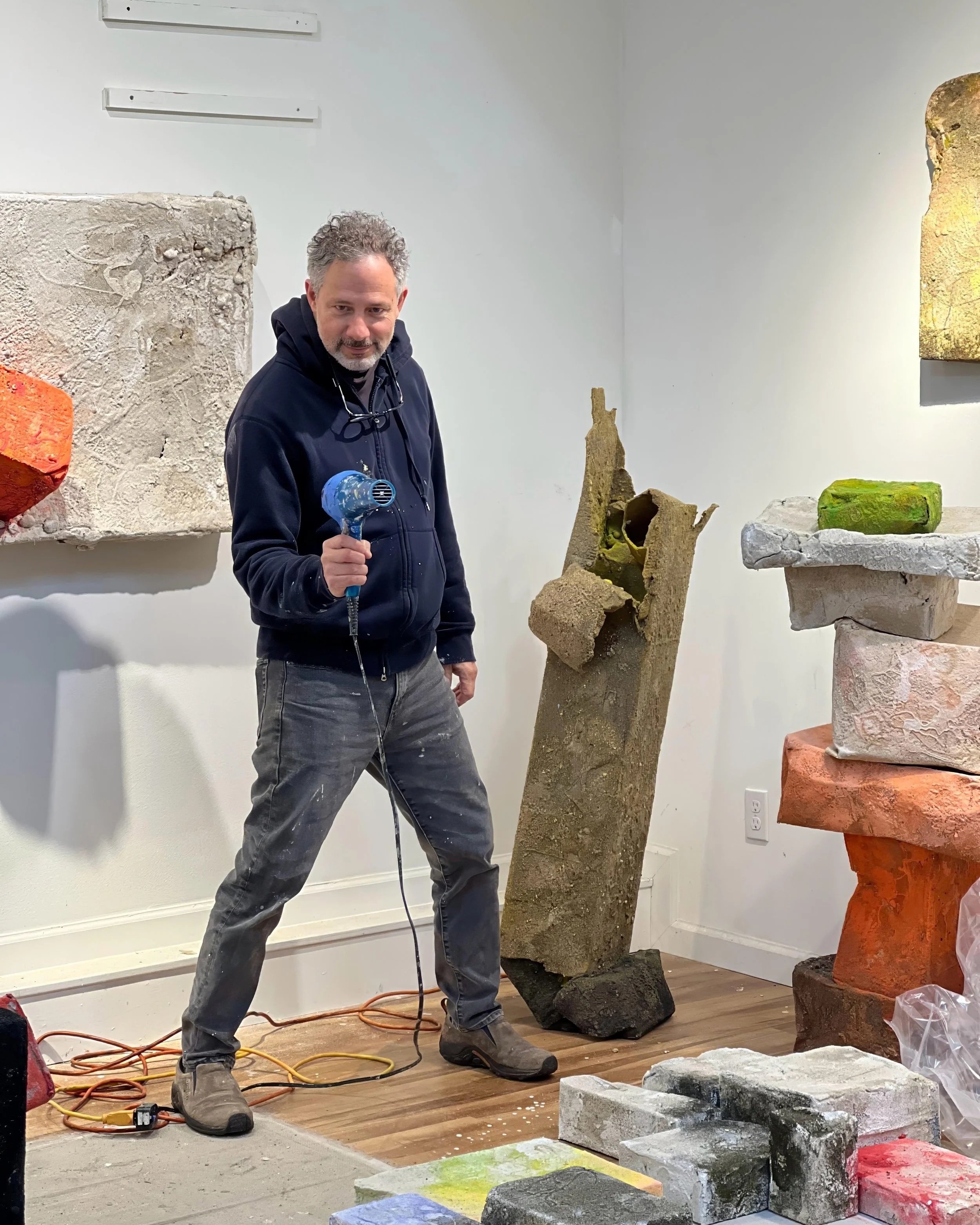 Sculptor and artist Mark Van Wagner, holding a blow dryer and standing in an art studio or gallery assembling his various large, textured sand sculptures and blocks on the floor and shelves.