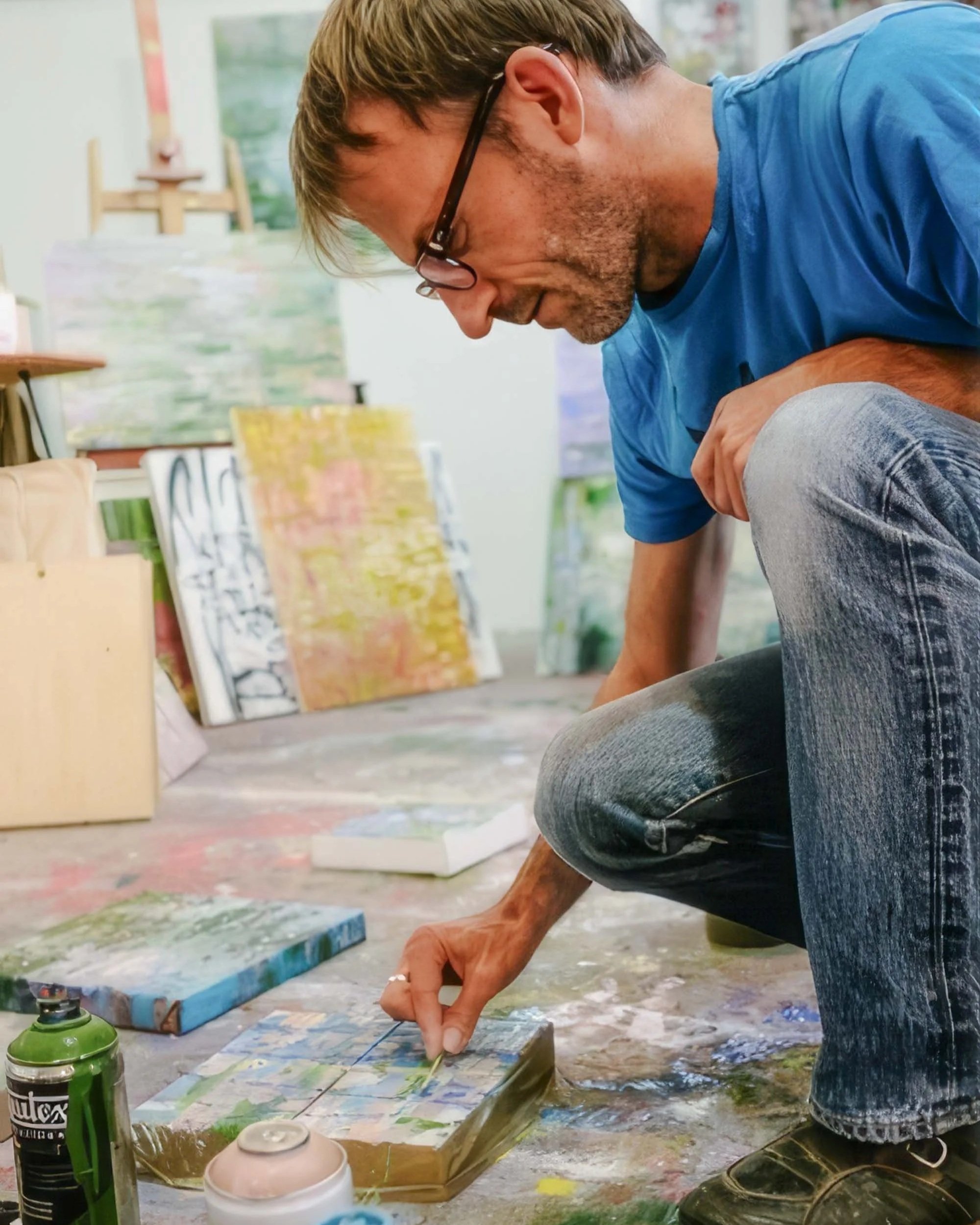 Close-up of artist Wayne Sleeth wearing glasses and a blue shirt, crouched down, painting on a canvas with a brush in an art studio. There are paint supplies and other artworks visible in the background.
