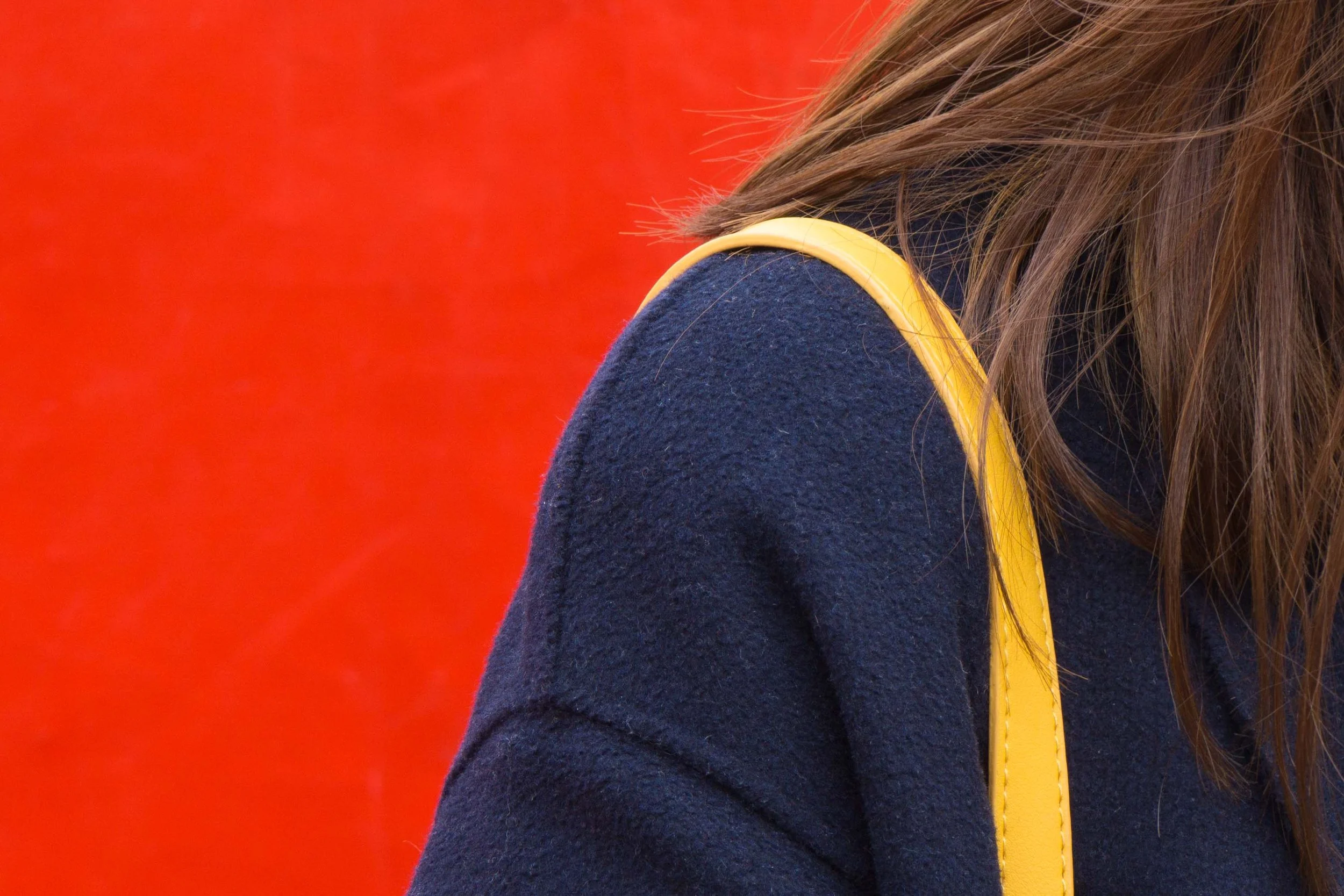 Abstract primary color photograph by artist Kimberly Poppe. Close-up of a woman's shoulder and side of her head, with long brown hair, wearing a navy blue coat with a yellow strap or handle against a red background.