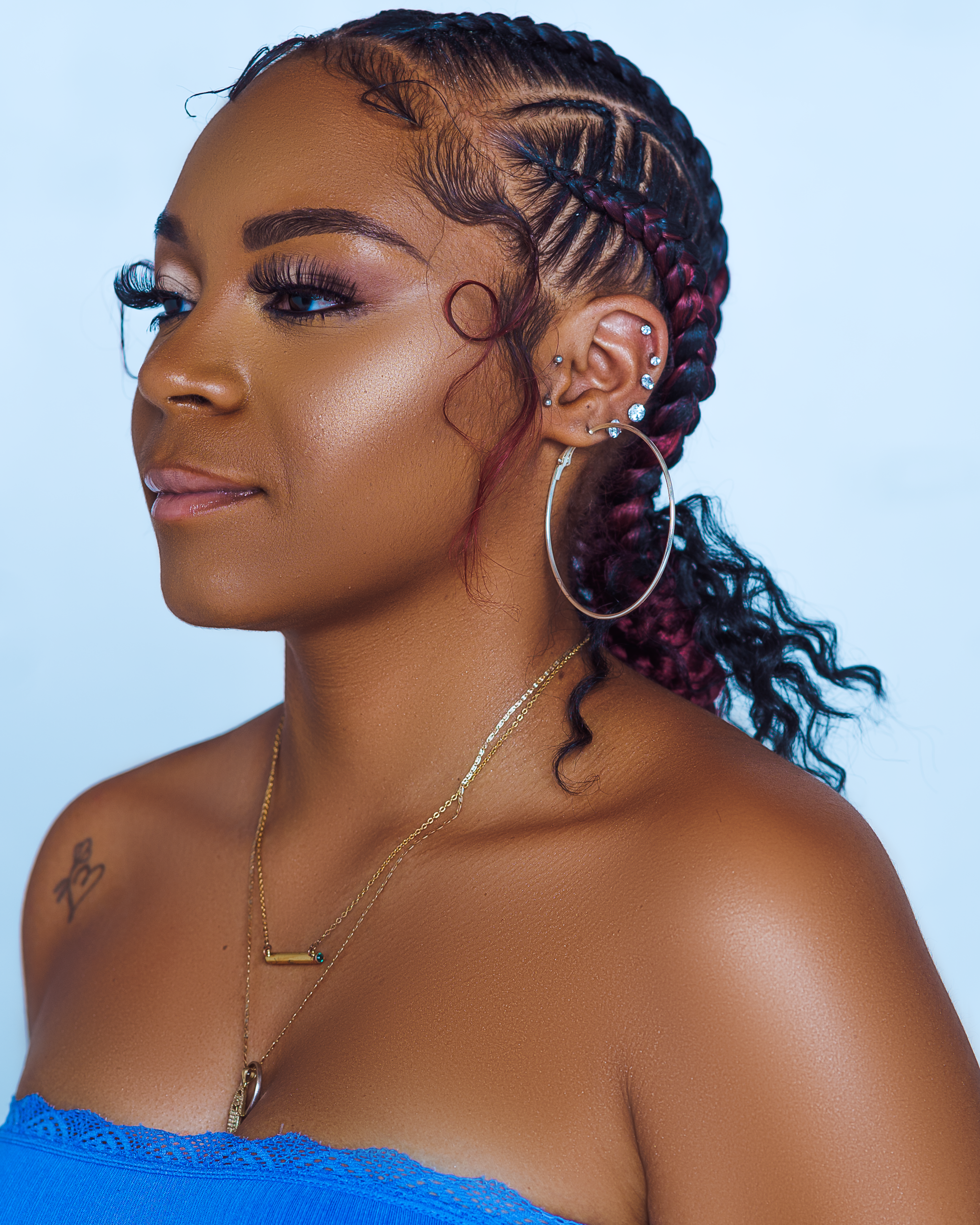 Close-up portrait of a woman with braided hair, multiple earrings, and layered necklaces, wearing a blue strapless top, against a plain background.