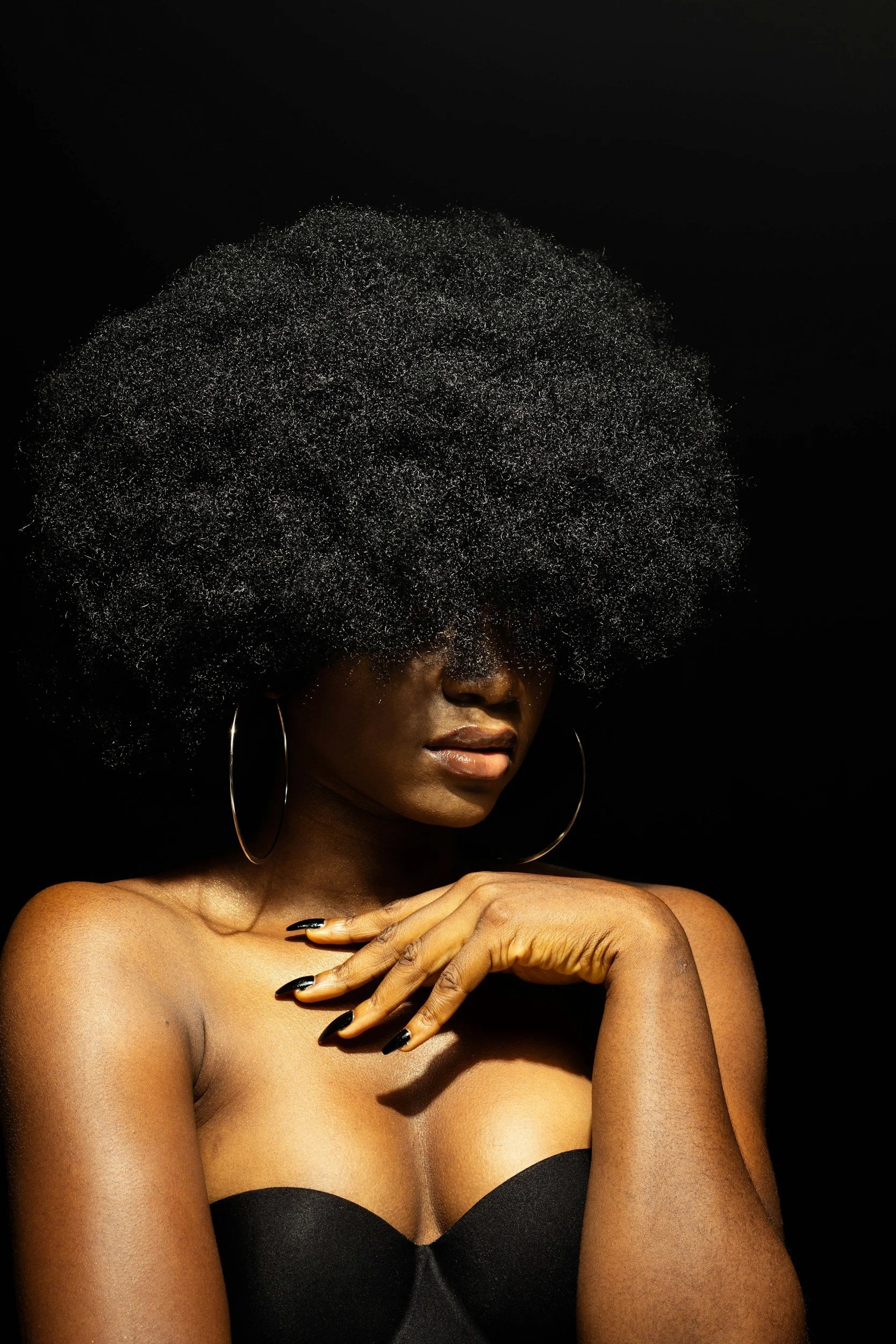 A woman with dark skin and curly black hair styled in an afro. She is wearing large hoop earrings, a strapless black top, and has black nail polish. Her hand is placed near her neck, and she is posing against a dark background.