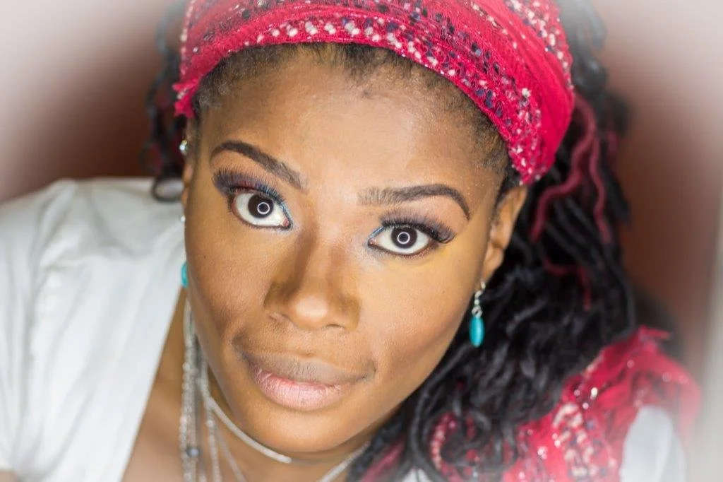 Close-up portrait of a woman with dark skin, wearing a red headband and jewelry, looking directly at the camera.