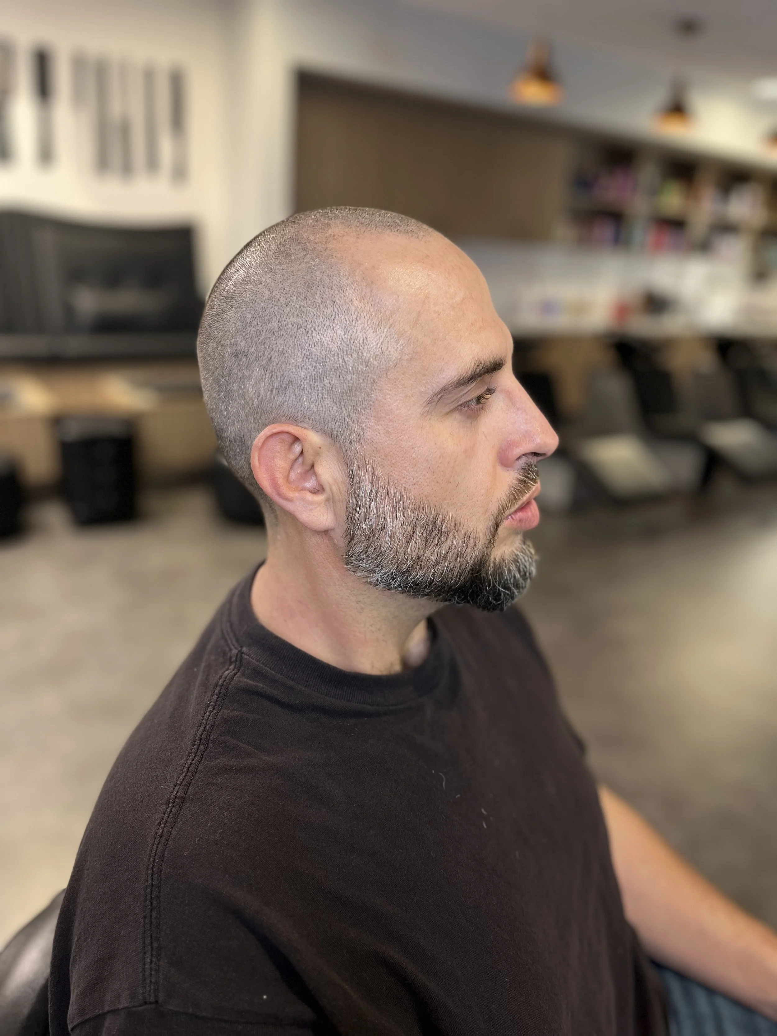 Side profile of a man with a shaved head and beard, wearing a black T-shirt, sitting in a room with blurred background including chairs, shelves, and lighting.