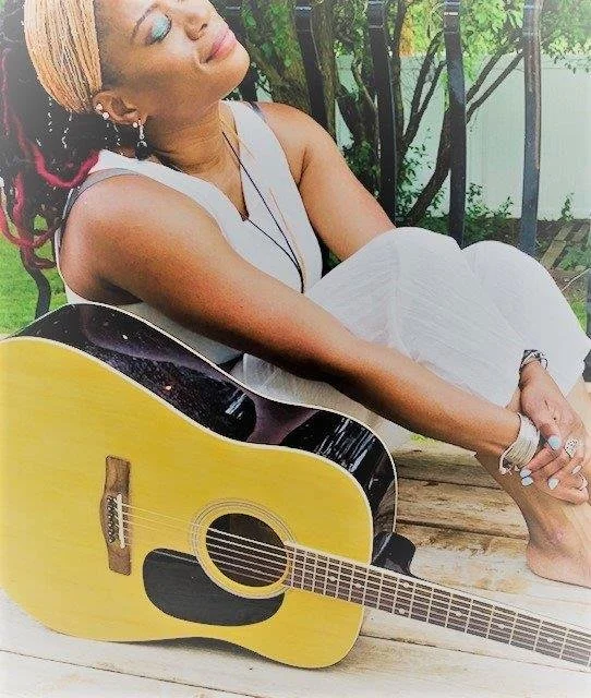 A woman sitting outdoors on a wooden surface with a yellow acoustic guitar, wearing a white dress, and smiling with eyes closed.