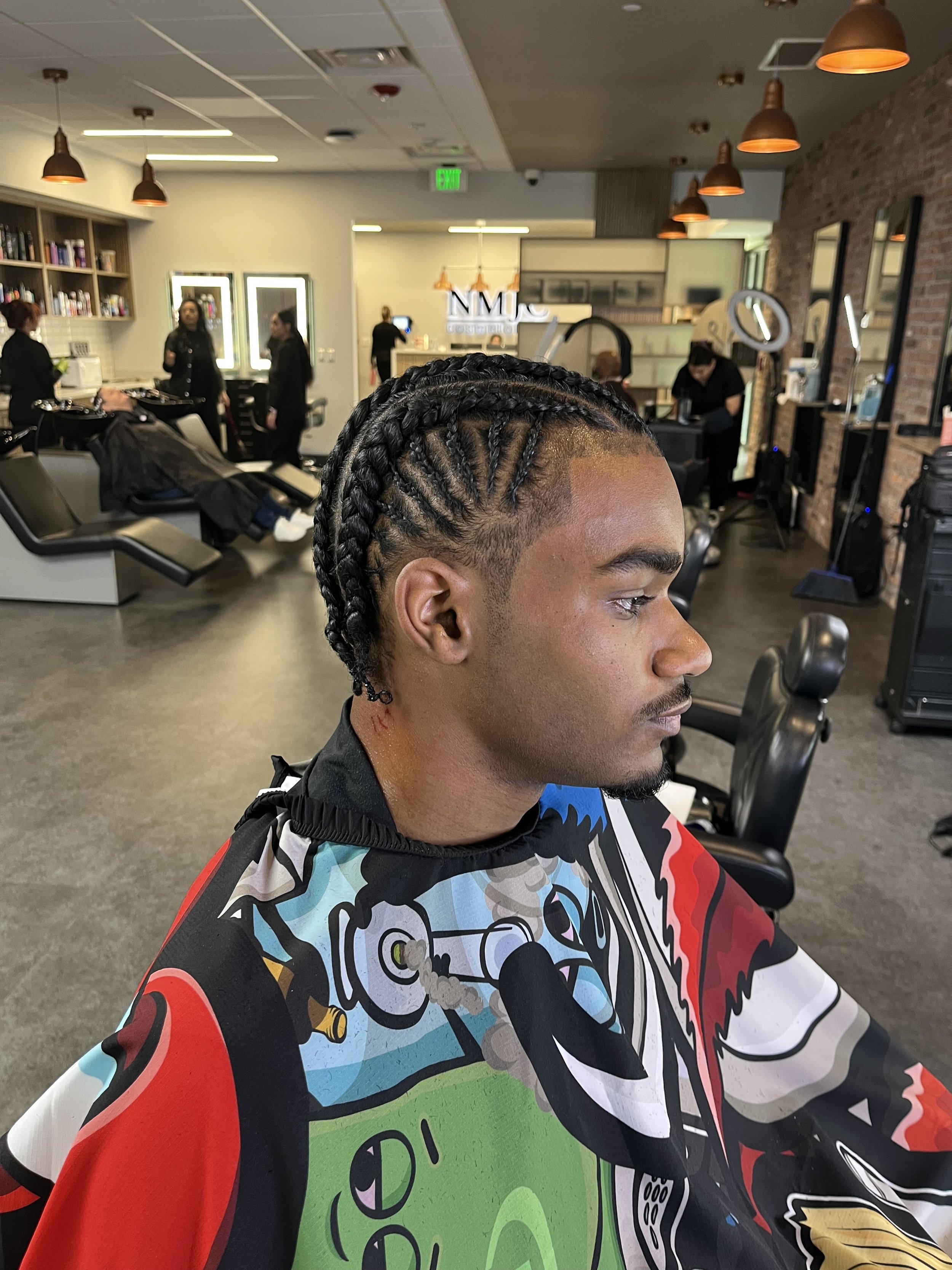 A man with braided hair and a colorful barber cape sitting in a hair salon or barbershop.