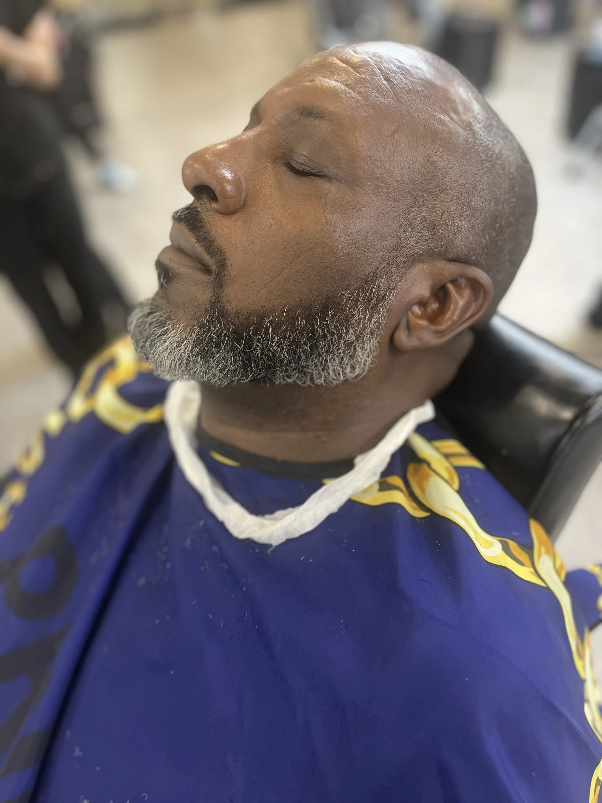 Close-up of a man with a shaved head and a gray beard, relaxing with eyes closed at a barbershop or salon.