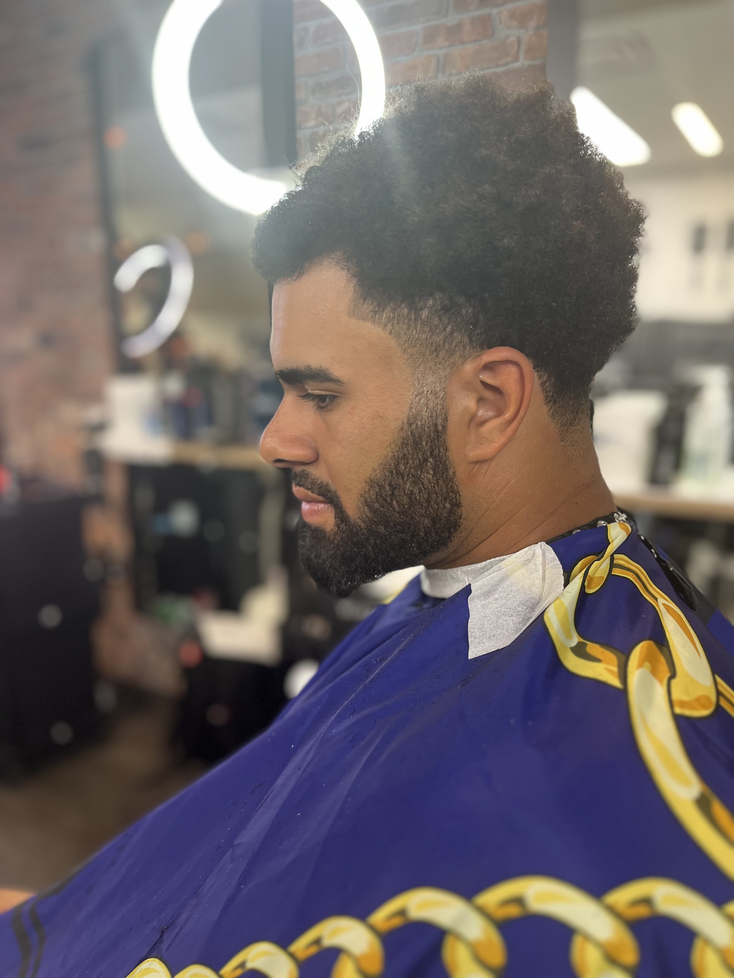 A man with a well-groomed beard and curly hair sitting in a barber shop, wearing a barber cape with a yellow chain pattern, as he waits for a haircut.
