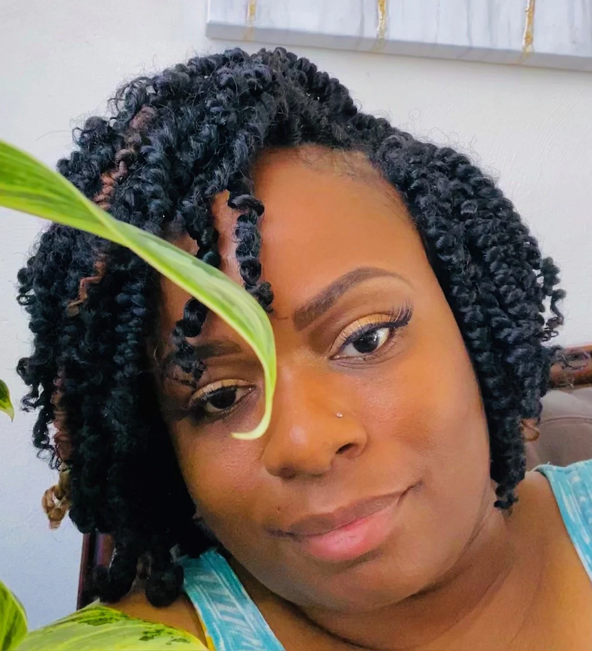Close-up of a woman with styled curly hair, wearing a blue striped top, with a leafy plant partially covering her face and a framed artwork in the background.