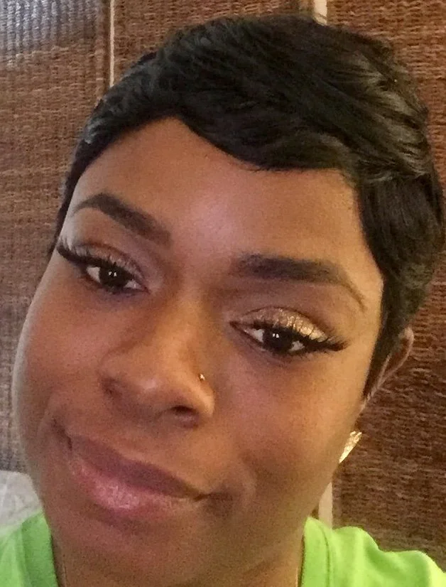 Close-up of a woman with short black hair, gold eye makeup, and a nose piercing, smiling with a wooden background.
