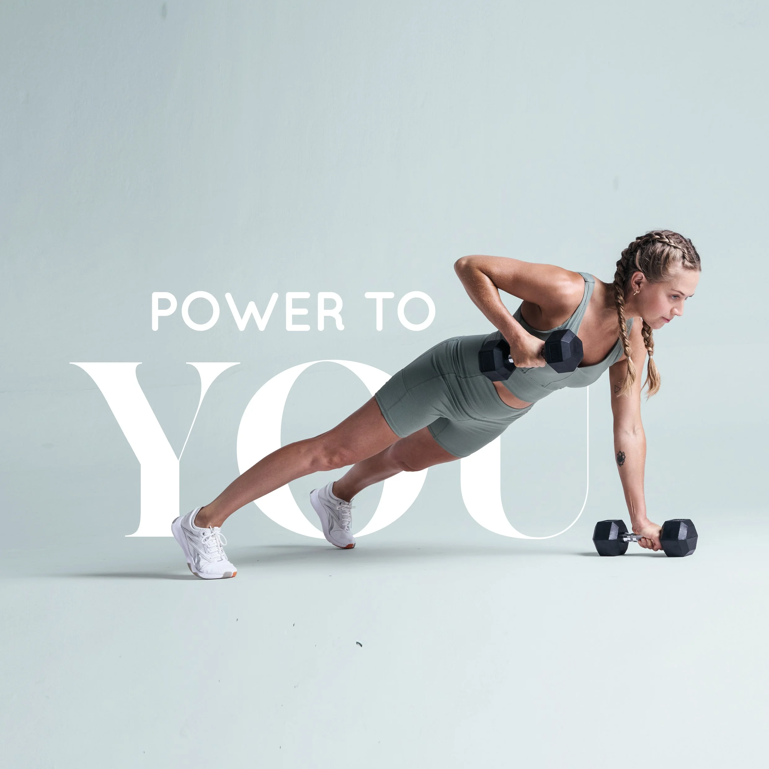 A woman in athletic clothing and braided hair is performing a plank exercise while holding a dumbbell in each hand against a plain light-colored background.