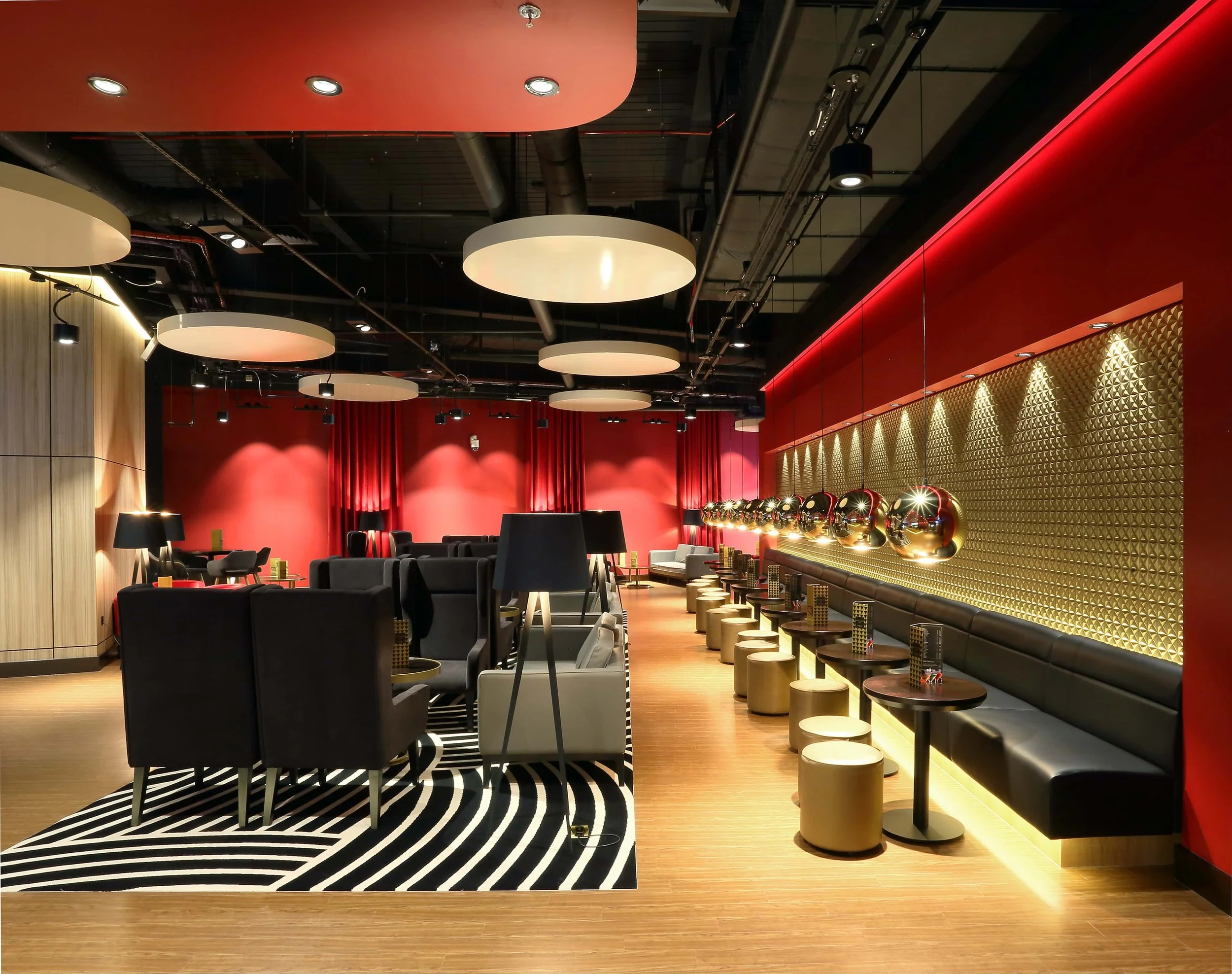 Interior of a modern lounge or waiting area with black chairs, beige stools, gold decor accents, red walls, circular ceiling lights, and a black and white striped rug.