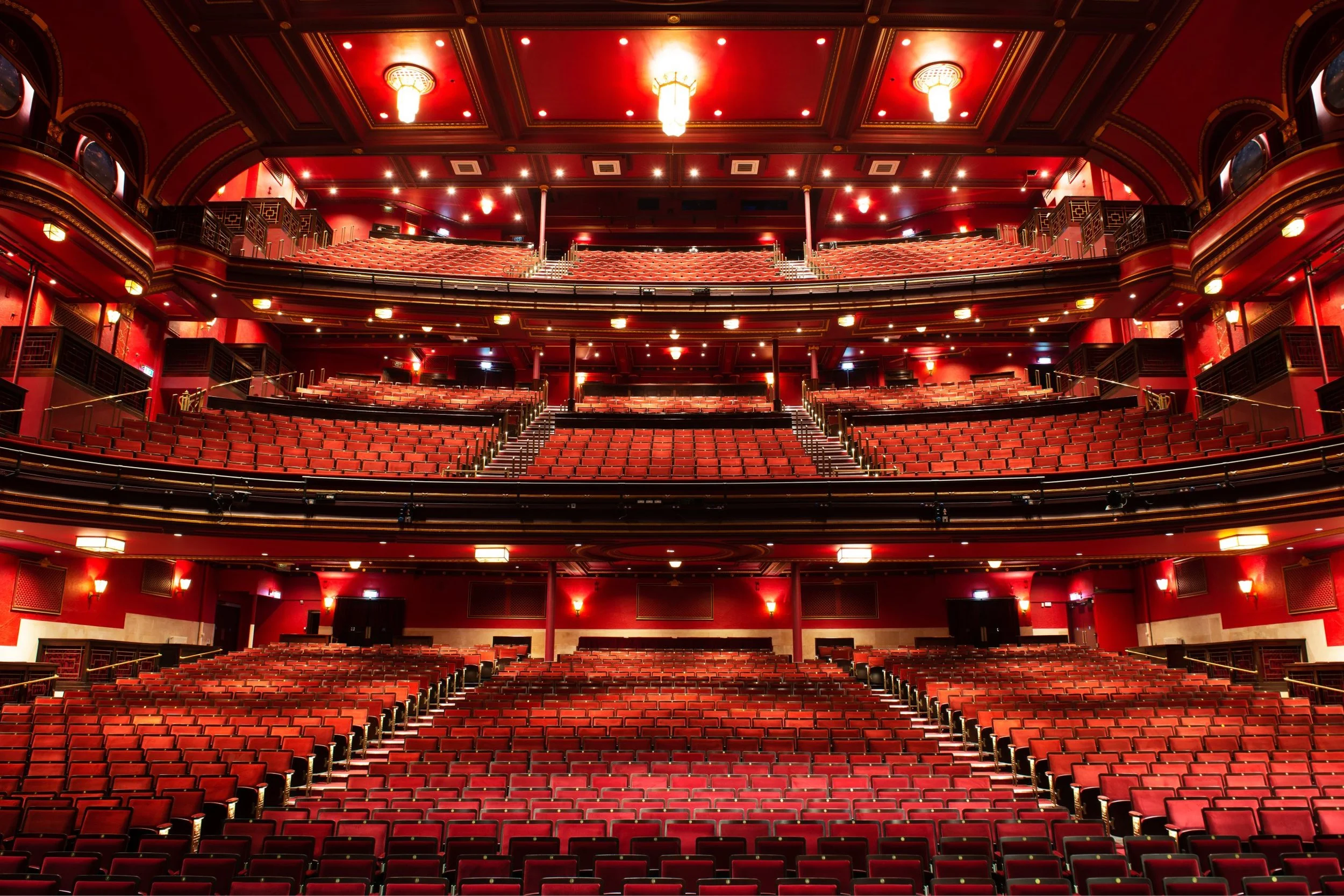 Inside of a grand theater with red walls and multiple levels of red seats, chandeliers hanging from the ceiling, and ornate details.