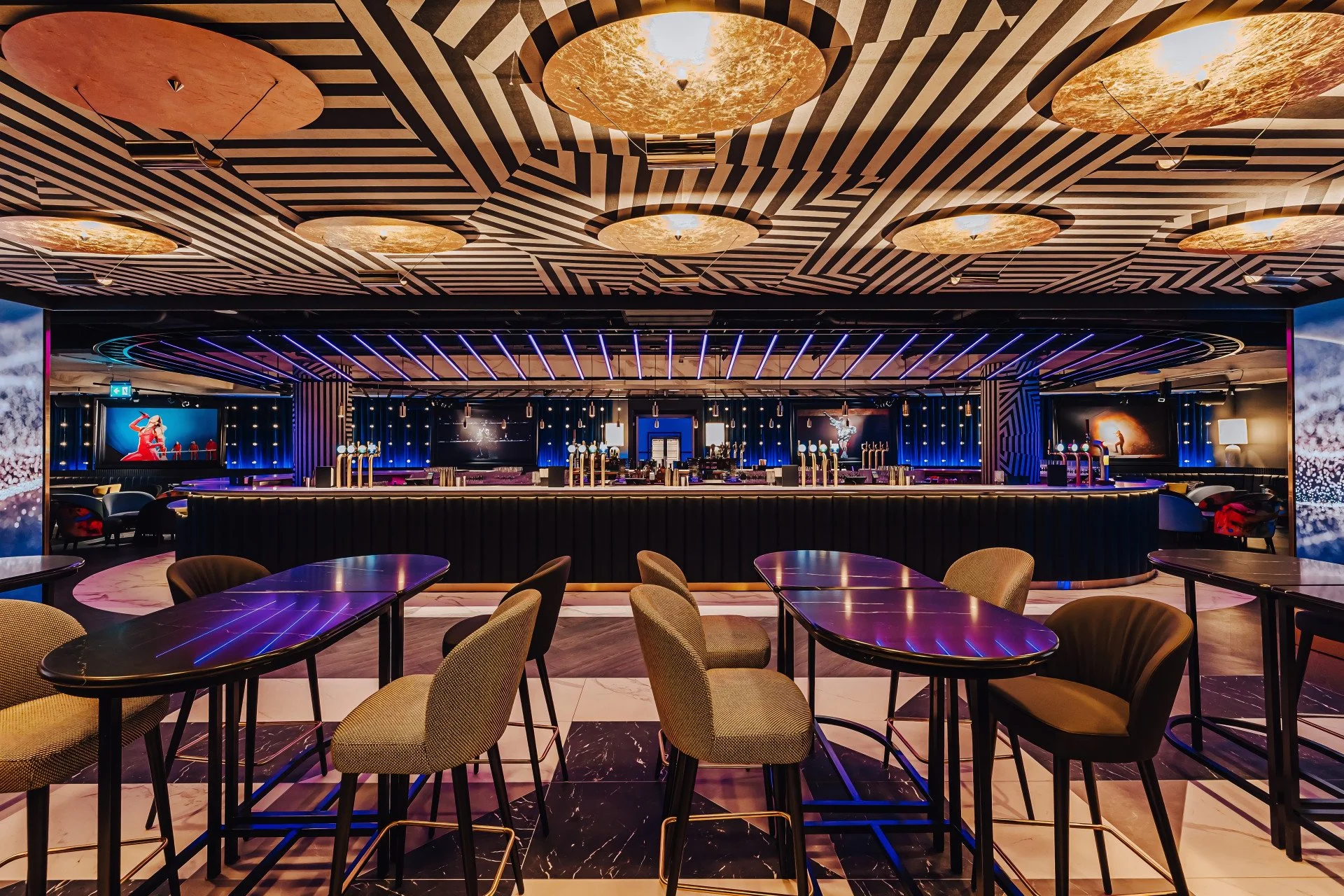 A modern, stylish bar with a geometric patterned ceiling featuring circular gold accents and purple lighting. The bar counter has several beer taps, and there are multiple screens and seating areas with tables and chairs in the foreground.
