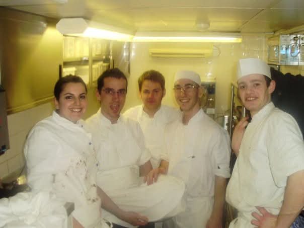 Group of five chefs in white uniforms smiling inside a commercial kitchen.