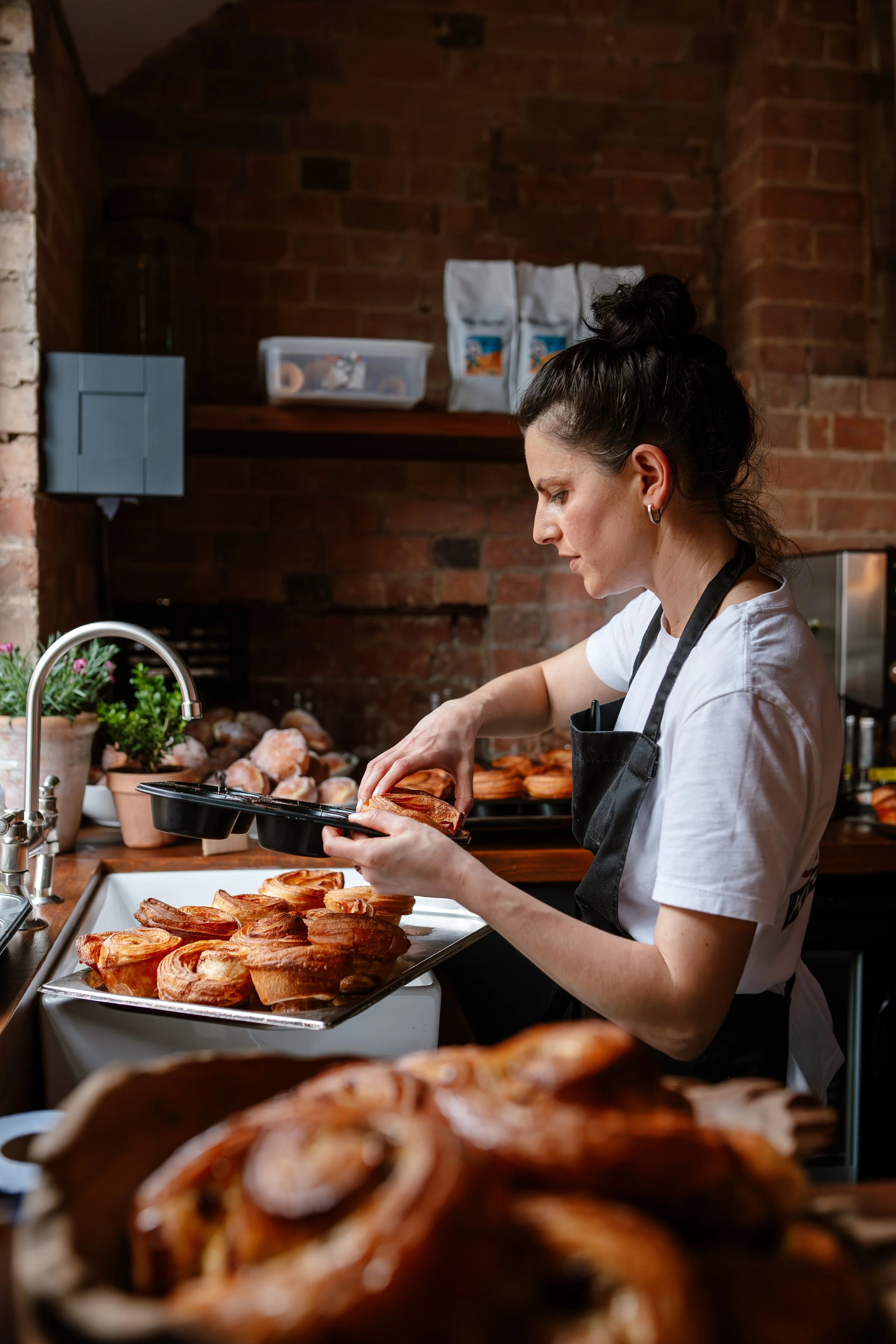 A woman with dark hair in a bun wearing a white shirt and black apron preparing baked goods in a rustic kitchen with brick walls. She is holding a tray of pastries and placing them on a white tray, with other baked goods visible in the background.