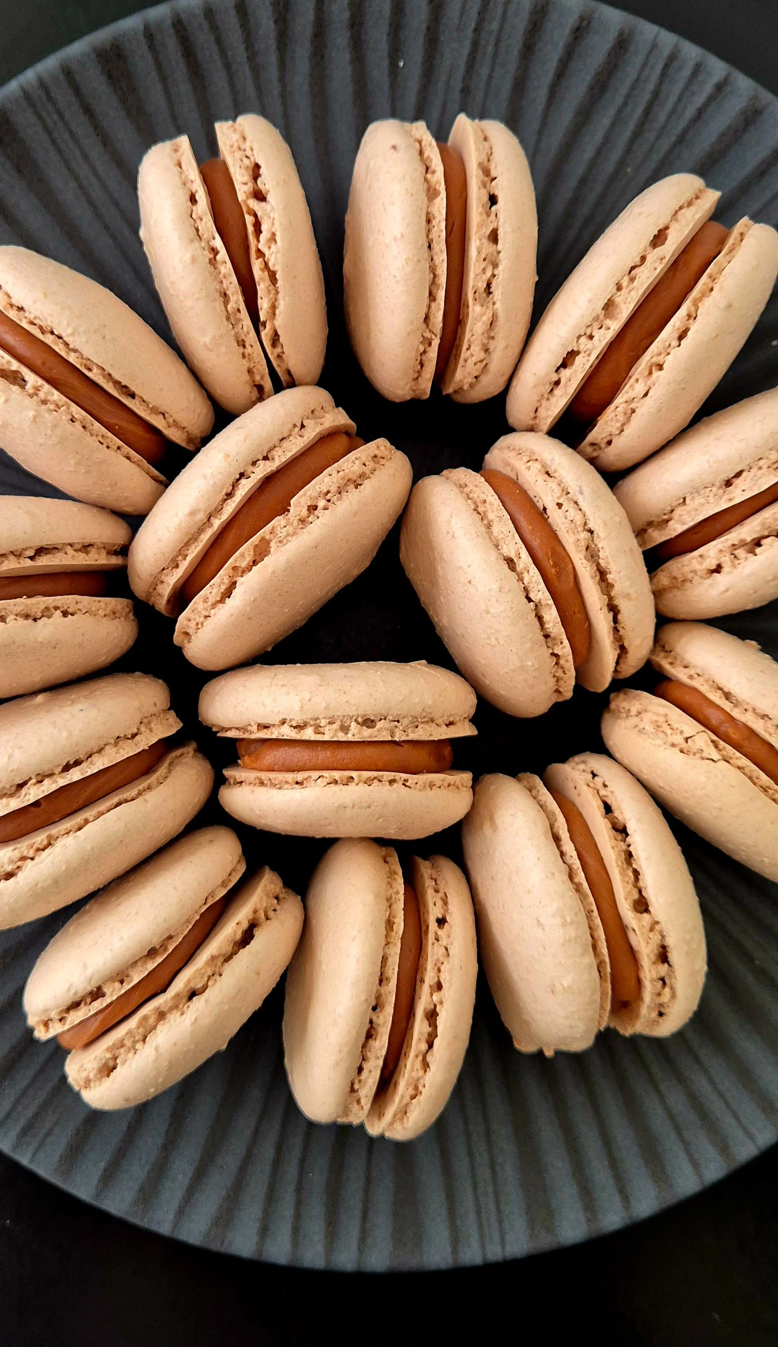 A black plate filled with beige macarons with caramel filling, arranged in a circular pattern.