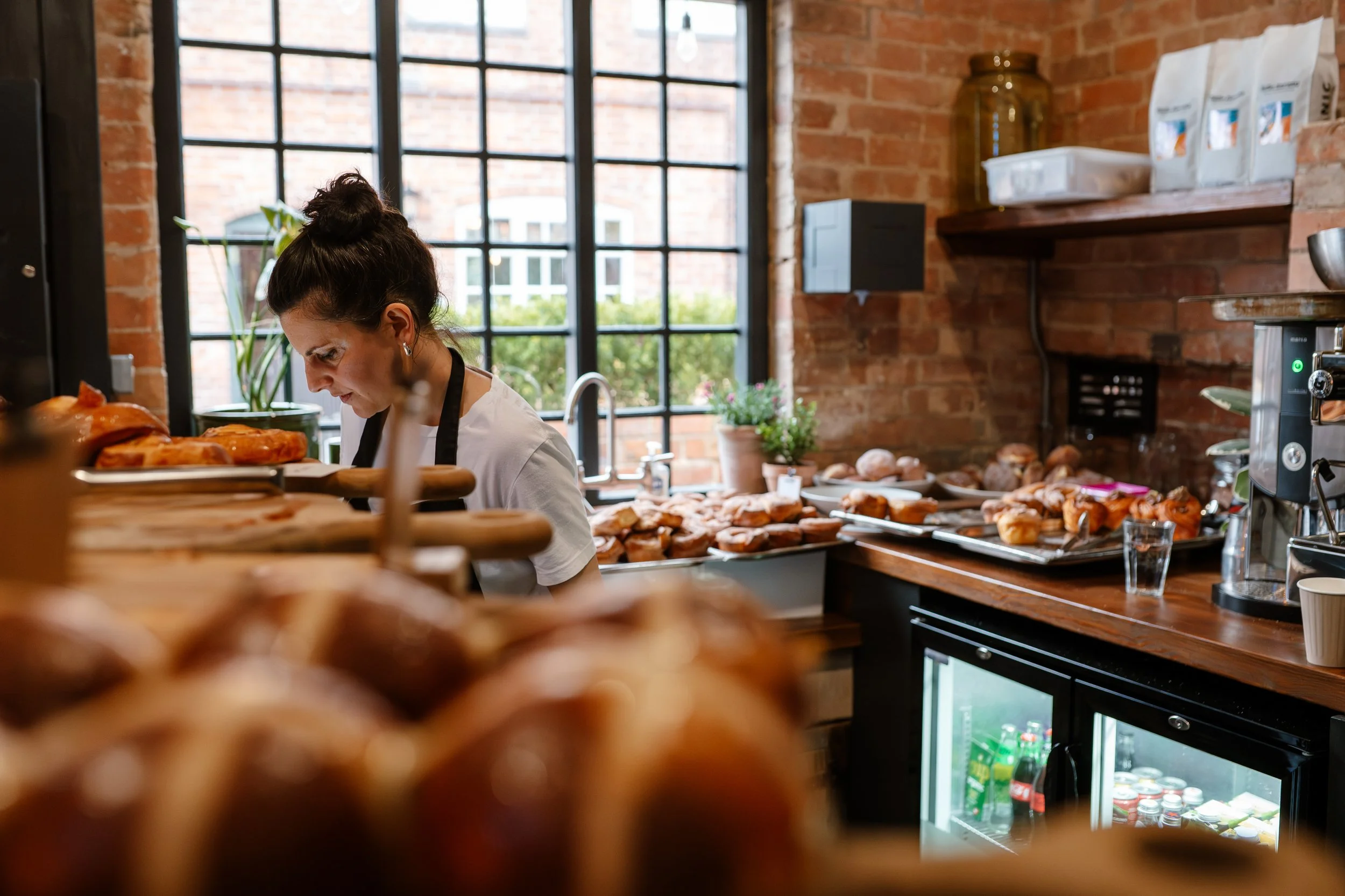 A woman with dark hair tied in a bun, wearing a white shirt and black apron, leaning over a table filled with baked goods inside a cozy bakery with brick walls and large windows.