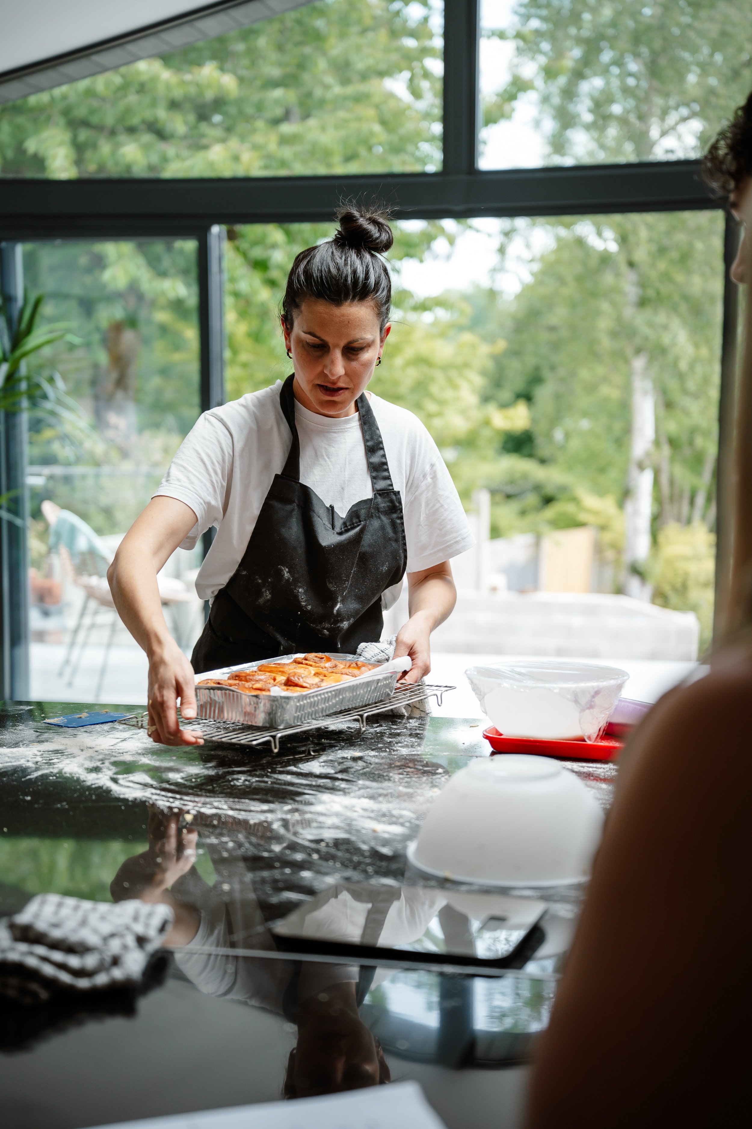 A woman wearing a black apron and white t-shirt is placing a tray of pizza into the oven of a kitchen with large windows, green trees visible outside.