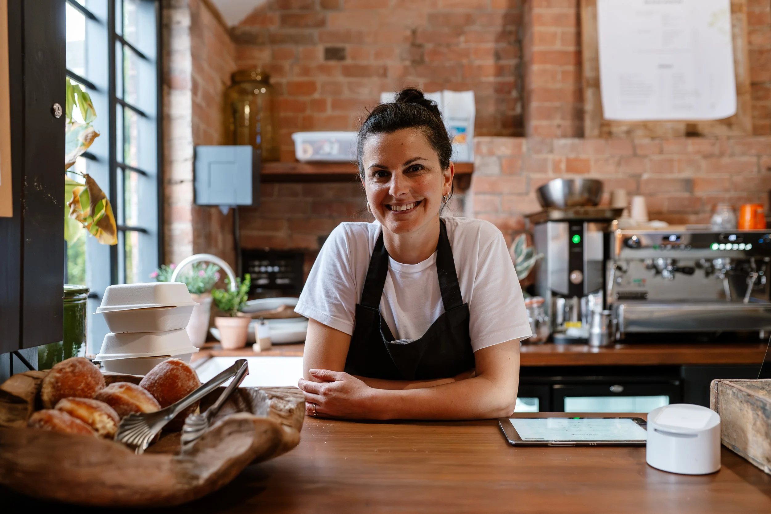 A smiling woman with dark hair tied up, wearing a white t-shirt and black apron, behind a wooden counter in a cafe or bakery, with baked goods and kitchen equipment in the background.