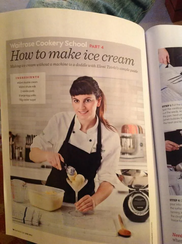 A woman in a white chef's coat and black apron demonstrating how to make ice cream, holding an ice cream scoop with ice cream in a kitchen.
