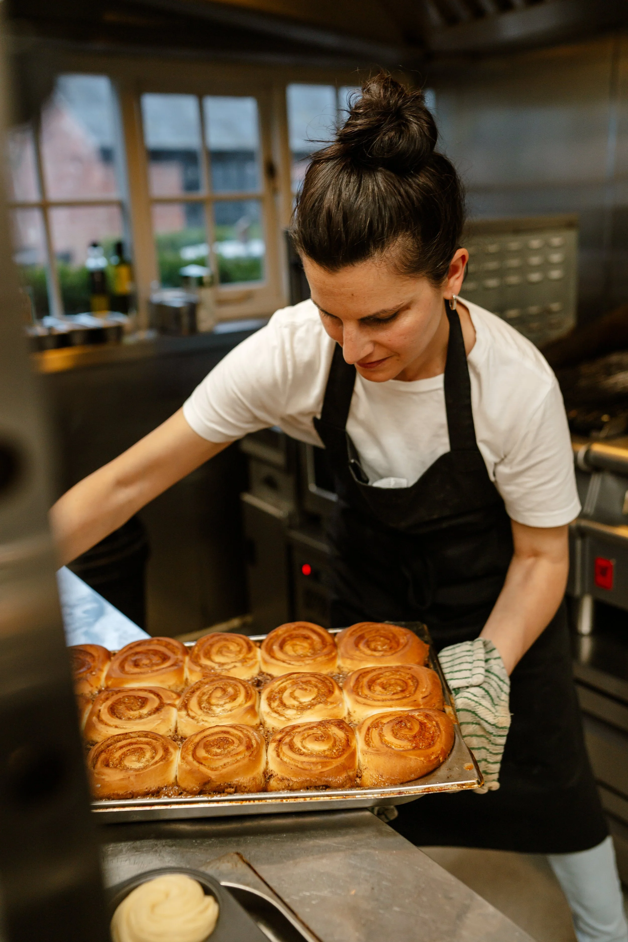 A woman with dark hair tied in a bun wearing a white t-shirt and black apron is holding a tray of freshly baked cinnamon rolls in a kitchen.