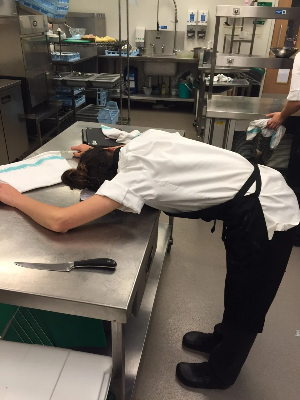 A chef is leaning over a stainless steel kitchen counter with their head resting on their arms, appearing exhausted or overwhelmed. They are wearing a white chef's jacket and a black apron. A large kitchen knife is on the counter nearby, and an arm of another person holding a dish towel is visible in the background.