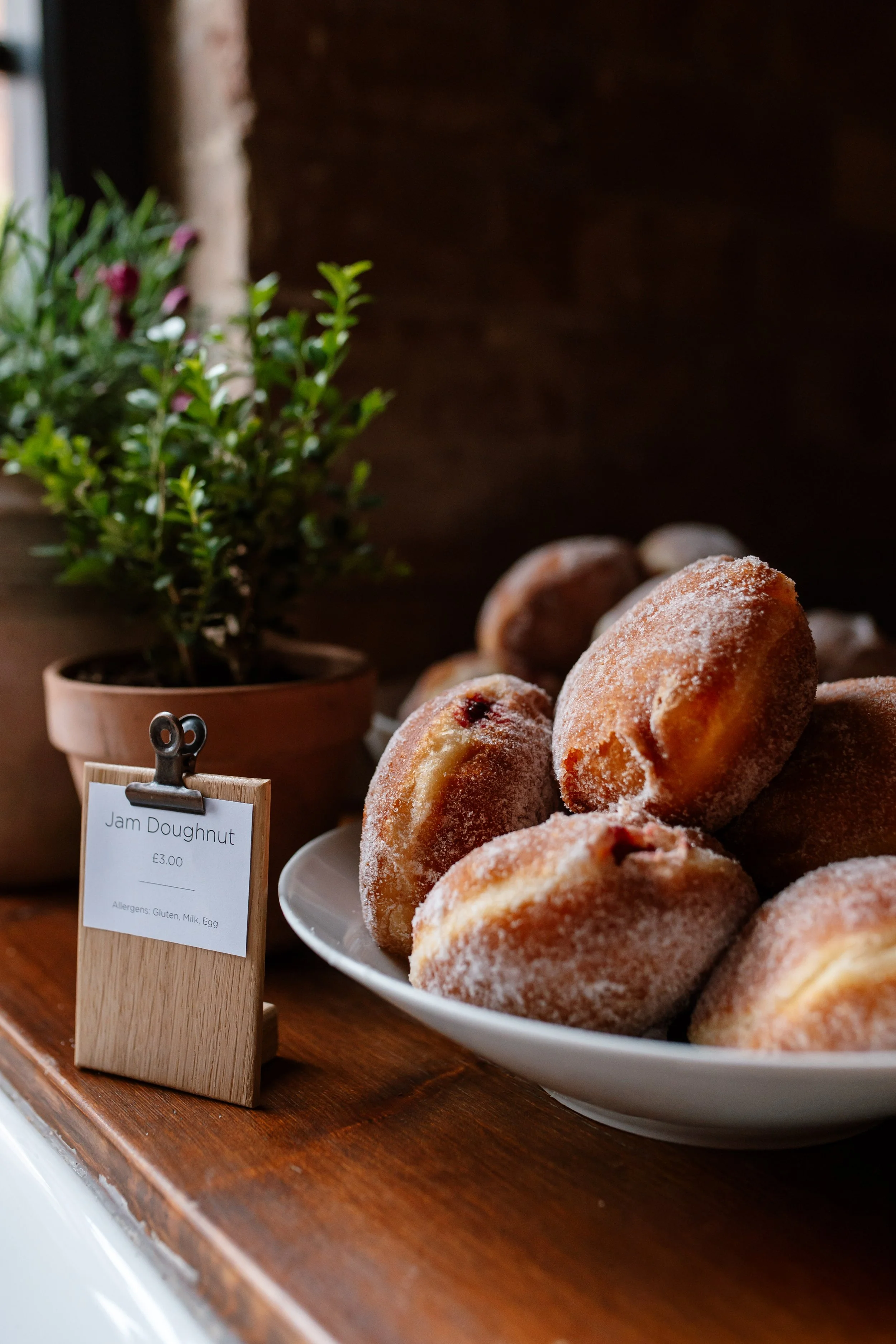 Plate of jam doughnuts coated with sugar, in front of a potted plant with green leaves and pink flowers, on a wooden surface with a small stand holding a sign that reads, 'Jam Doughnut, £3.00, Allergens: Gluten, Milk, Egg'.