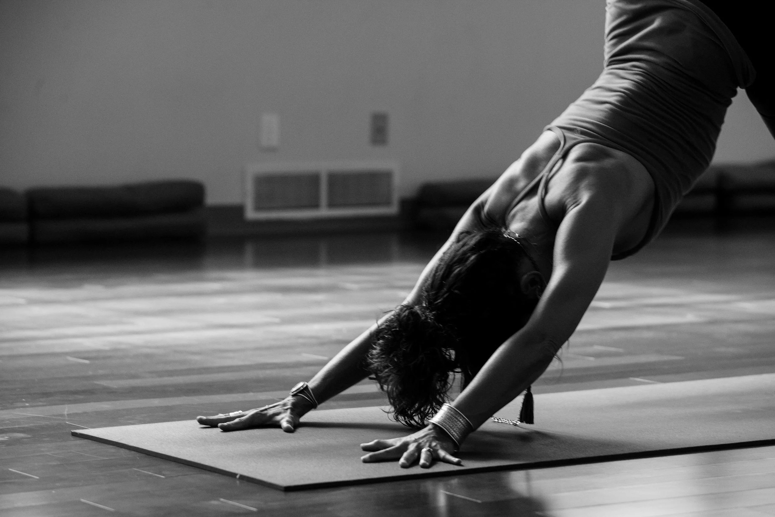 A woman practicing yoga on a mat in a dimly lit room, leaning forward with her hands on the floor and her head down, in a resting position.