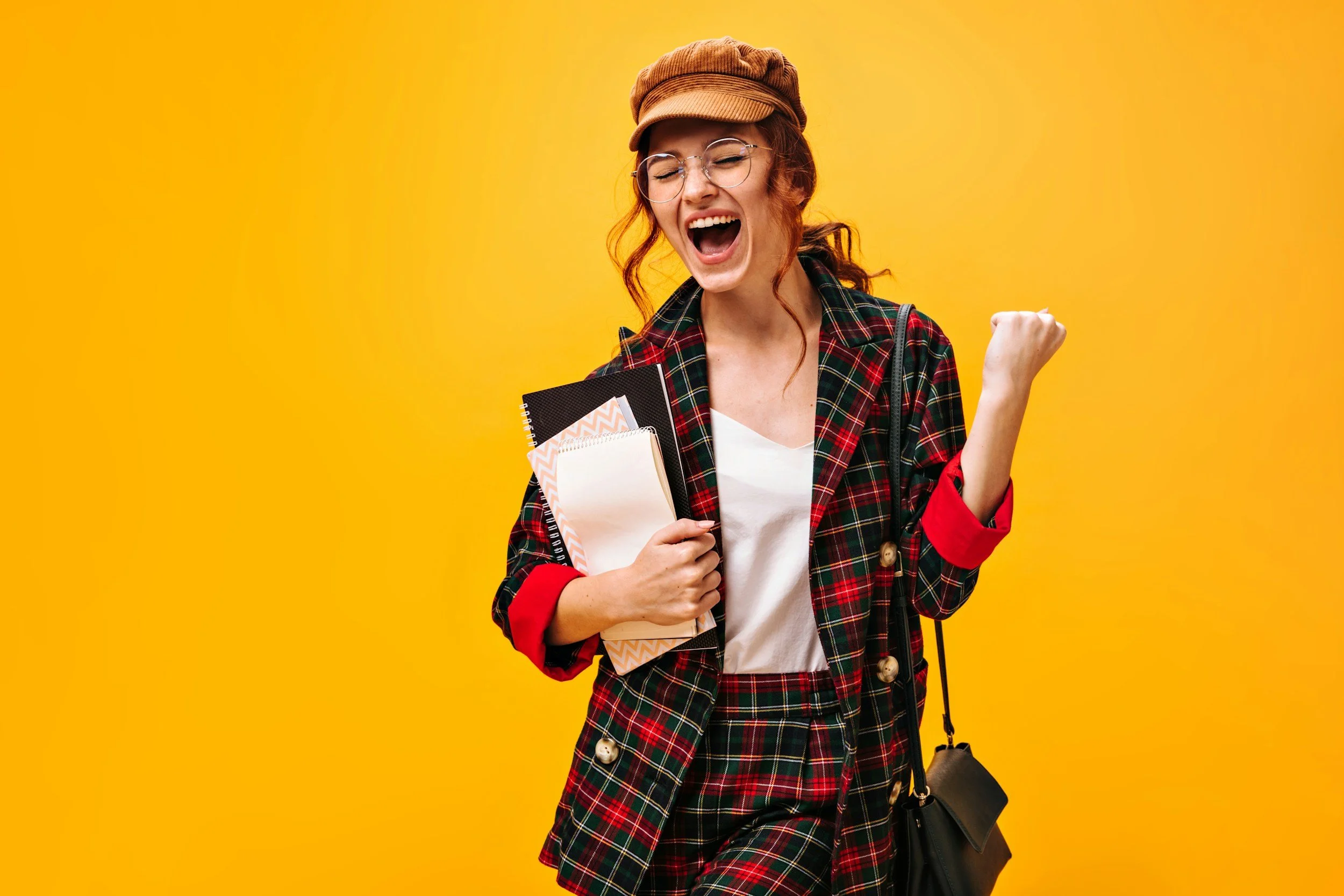 Junge Frau mit Brille, rotem kariertem Mantel, Mütze, hält Notizbuch, macht Siegerpose und freut sich vor gelbem Hintergrund.