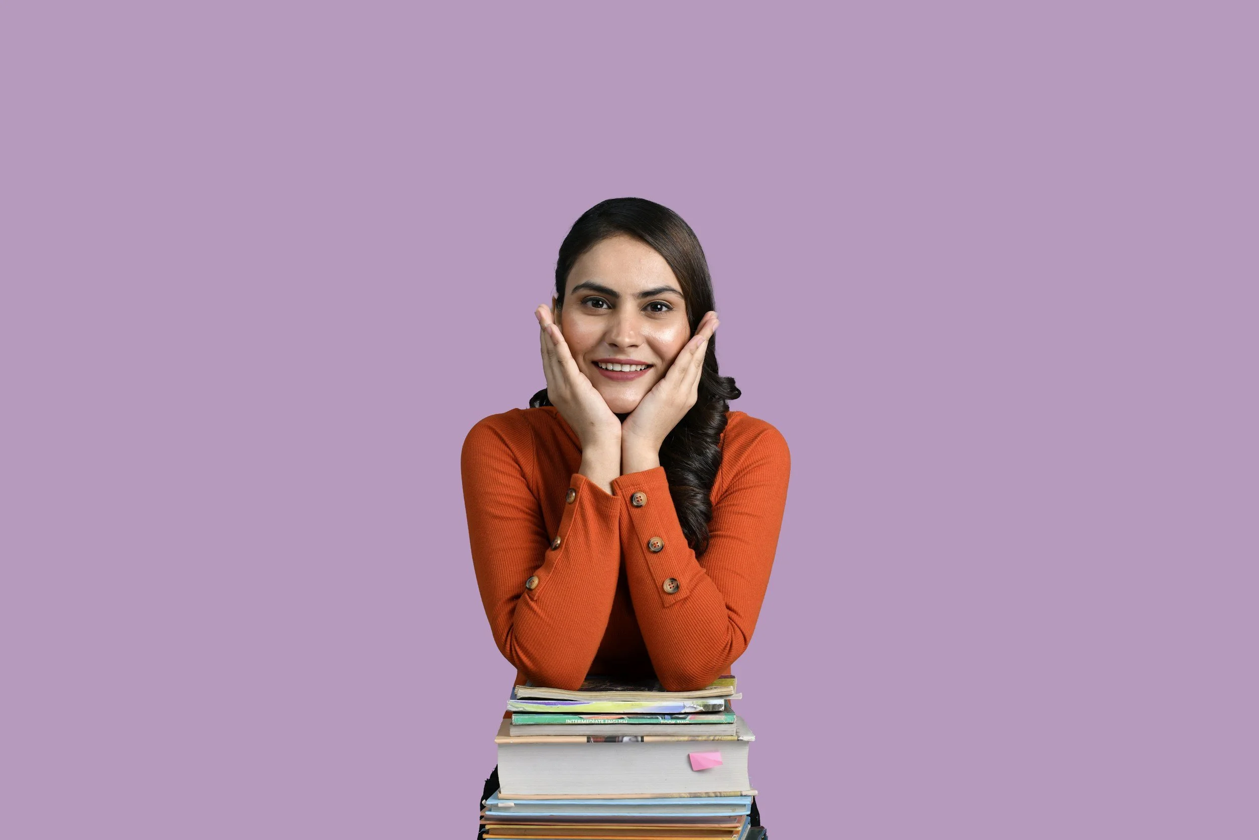 Junge Frau mit dunklen Haaren, lächelnd und die Hände an den Wangen, sitzt hinter einem Stapel Bücher auf lila Hintergrund.