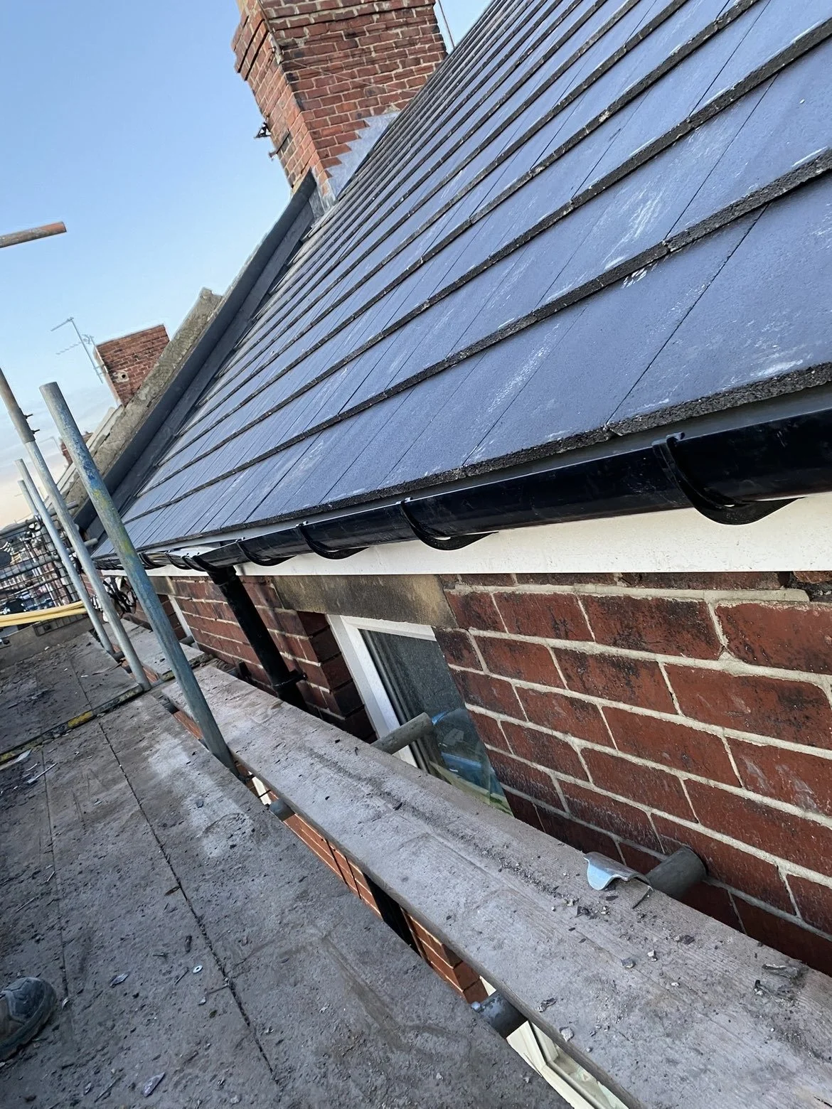 The image shows a close-up of a house's roof with black slates, a brick chimney, and a gutter system underneath. There is scaffolding with wooden planks on the side of the house, indicating ongoing construction or maintenance work.