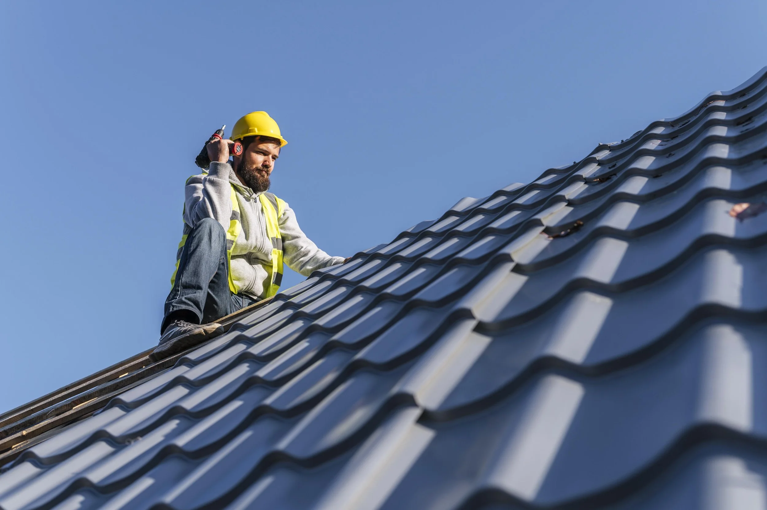 A construction worker wearing a yellow safety helmet and gray hoodie, kneeling on a roof with blue tiles, holding a power drill, with a clear blue sky in the background.
