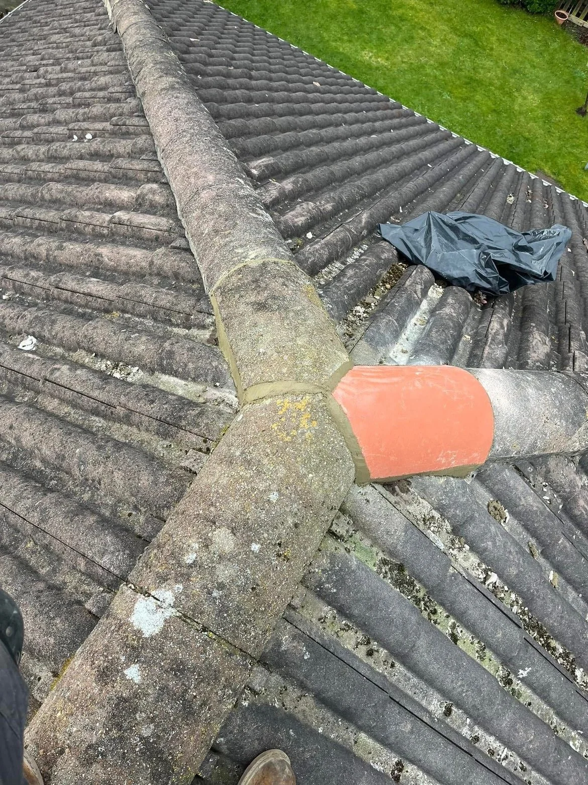 A roof with old, weathered tiles and a ridge that has some repair work with new mortar and a patch of orange sealant. A black plastic bag is placed on top of the tiles, and a grassy yard is visible in the background.