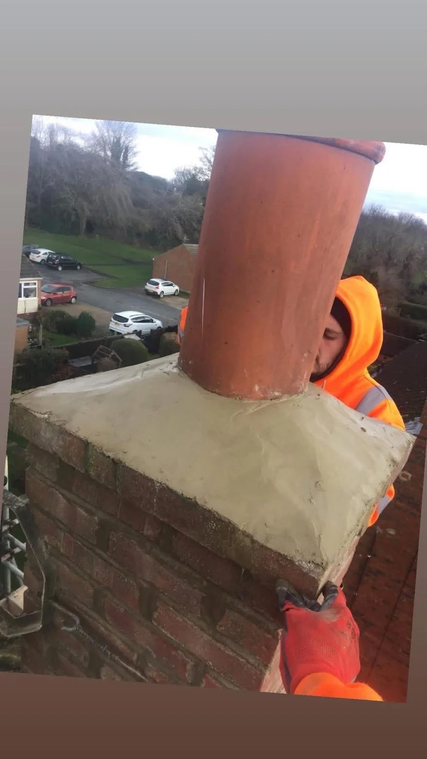 A worker in an orange hoodie and gloves is installing a chimney pipe on a brick chimney. The worker is holding the bricks of the chimney, and the photo is taken from an elevated angle.