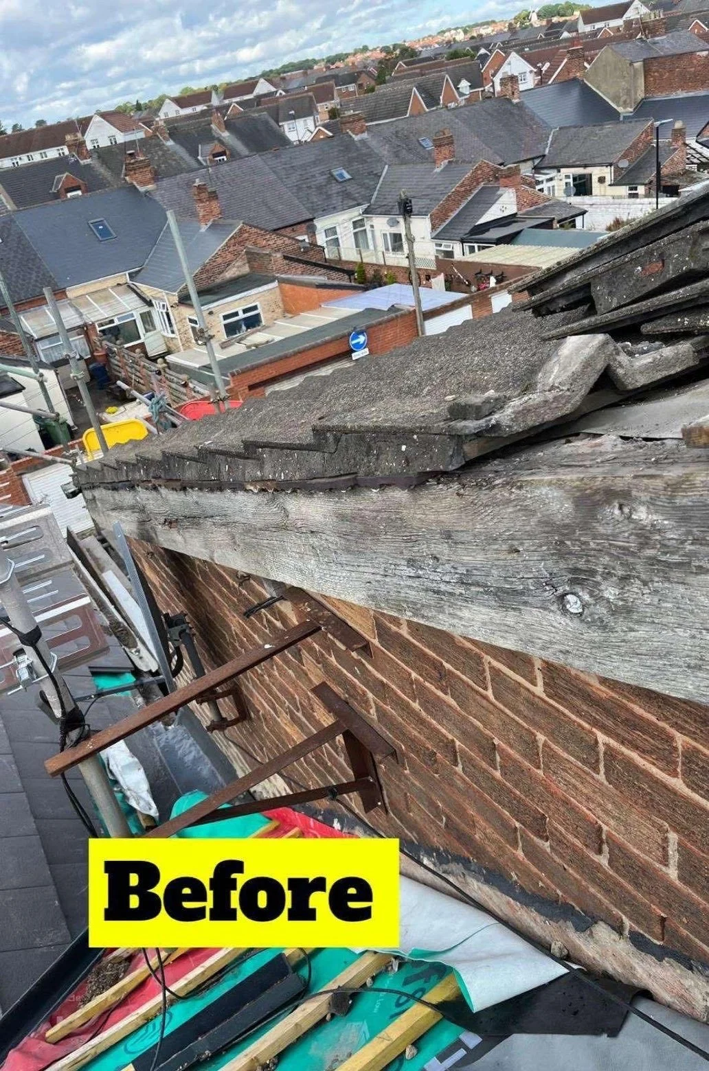 View of a rooftops and a neighborhood from a building with a damaged gutter and roof edge. The foreground shows a worn wooden beam and debris, and the background features multiple houses with chimneys and roof vents under a partly cloudy sky.