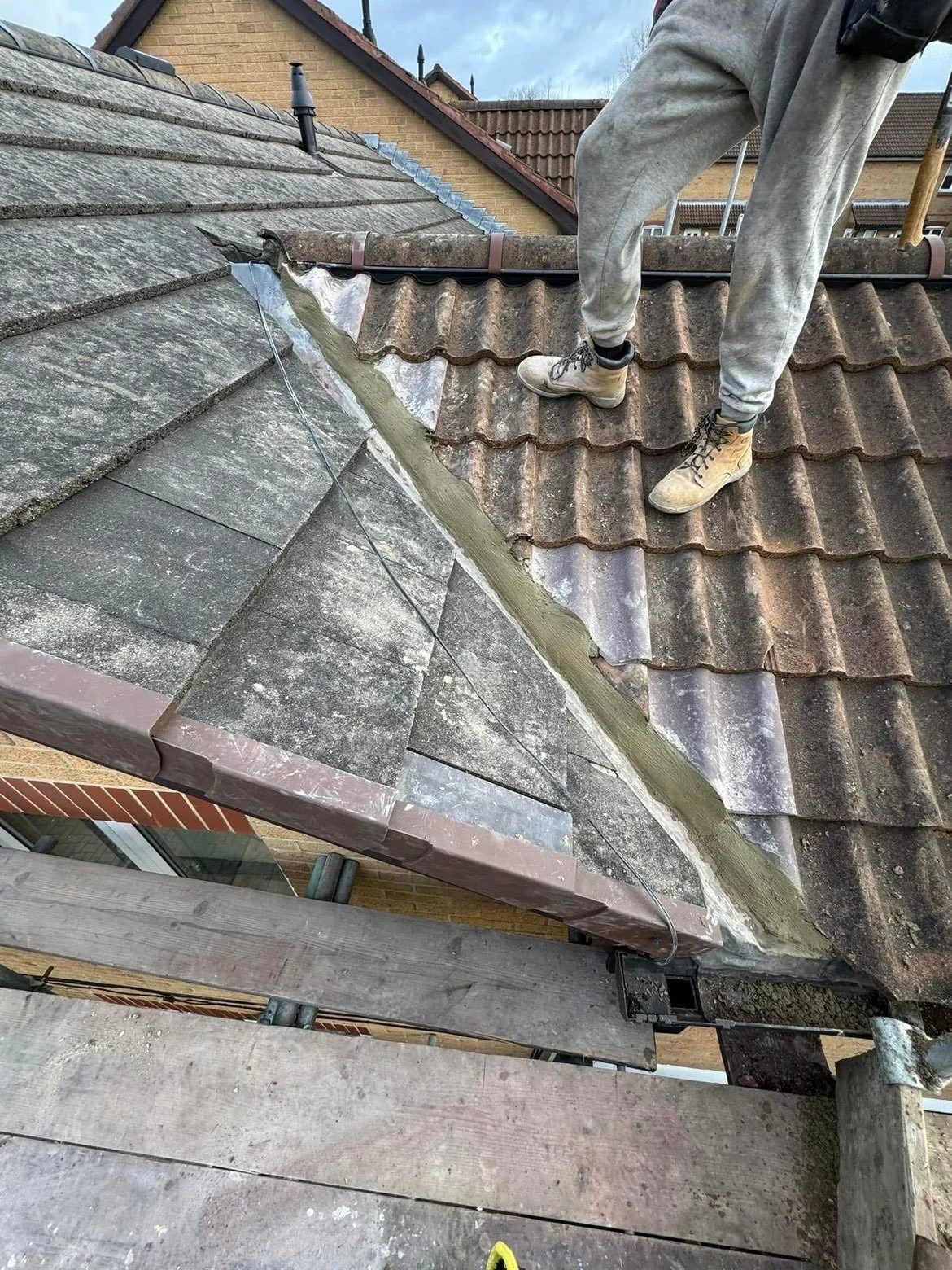 Worker on a roof repairing or installing flashing, standing on brown weathered tiles with scaffolding below.