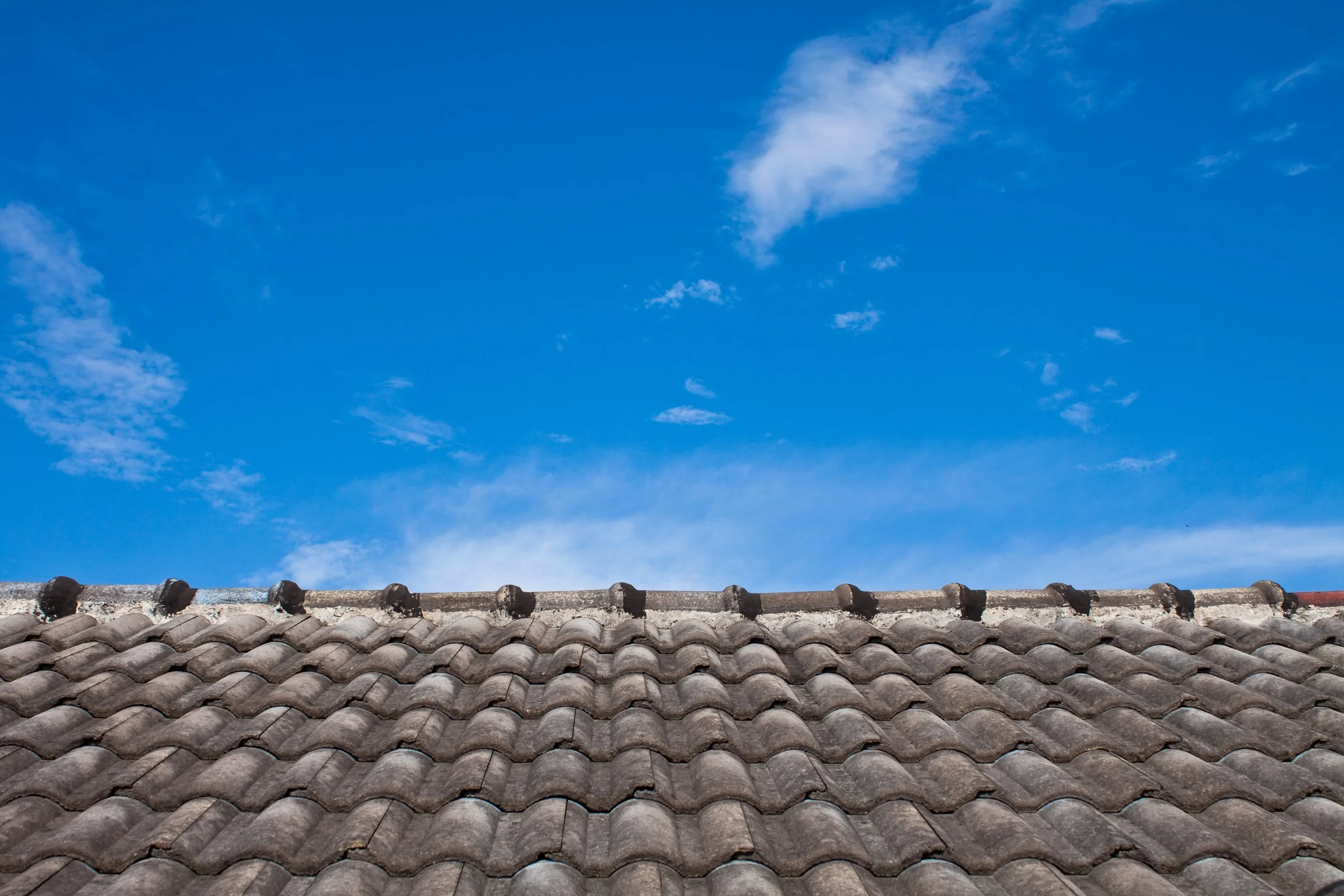 Photo of a blue sky with a few white clouds and a tiled roof at the bottom.