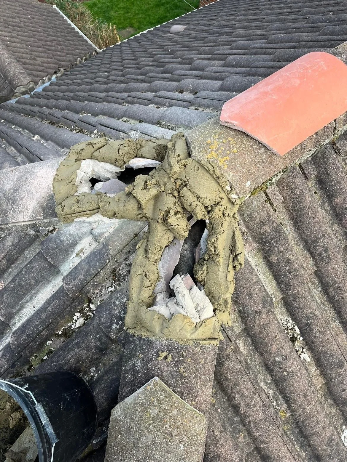The image shows a damaged chimney on a tiled roof with some of the tiles displaced and repair work in progress. There is a black bucket and a clear plastic container visible near the roof edge.