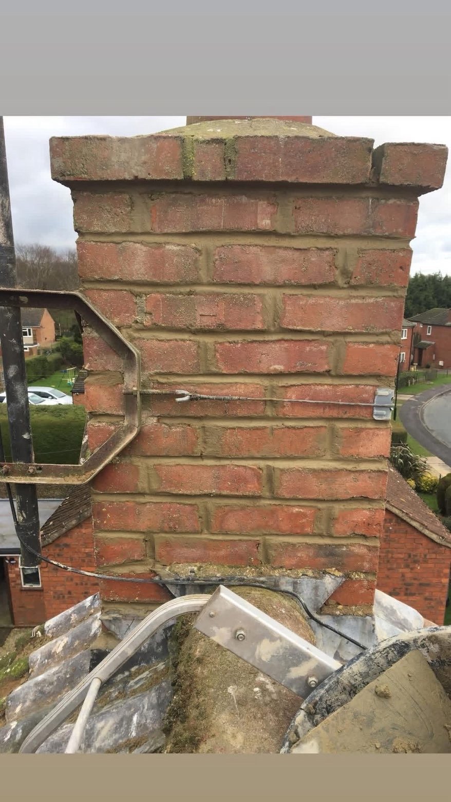 A brick chimney on a roof with a black metal frame to the side, some wires, and surrounding suburban houses and streets visible in the background.