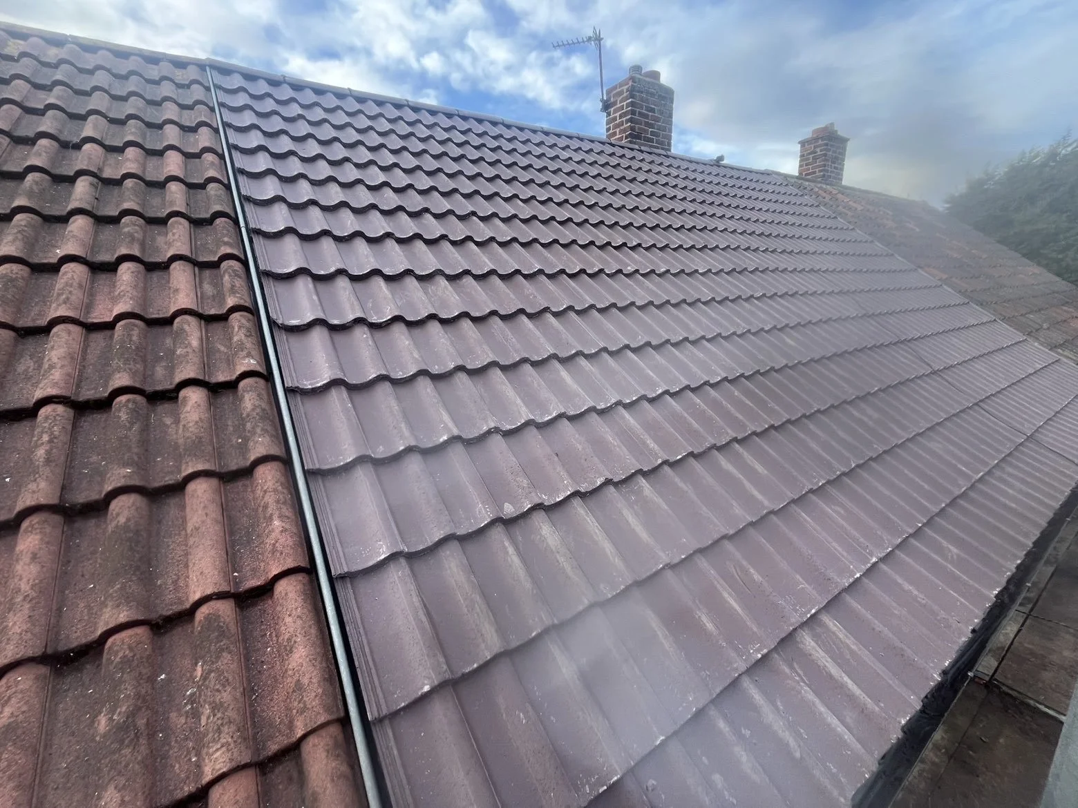 A roof with brown clay tiles, some of which are being replaced with new purple tiles. Two chimneys are visible in the background, along with an antenna and partly cloudy sky.