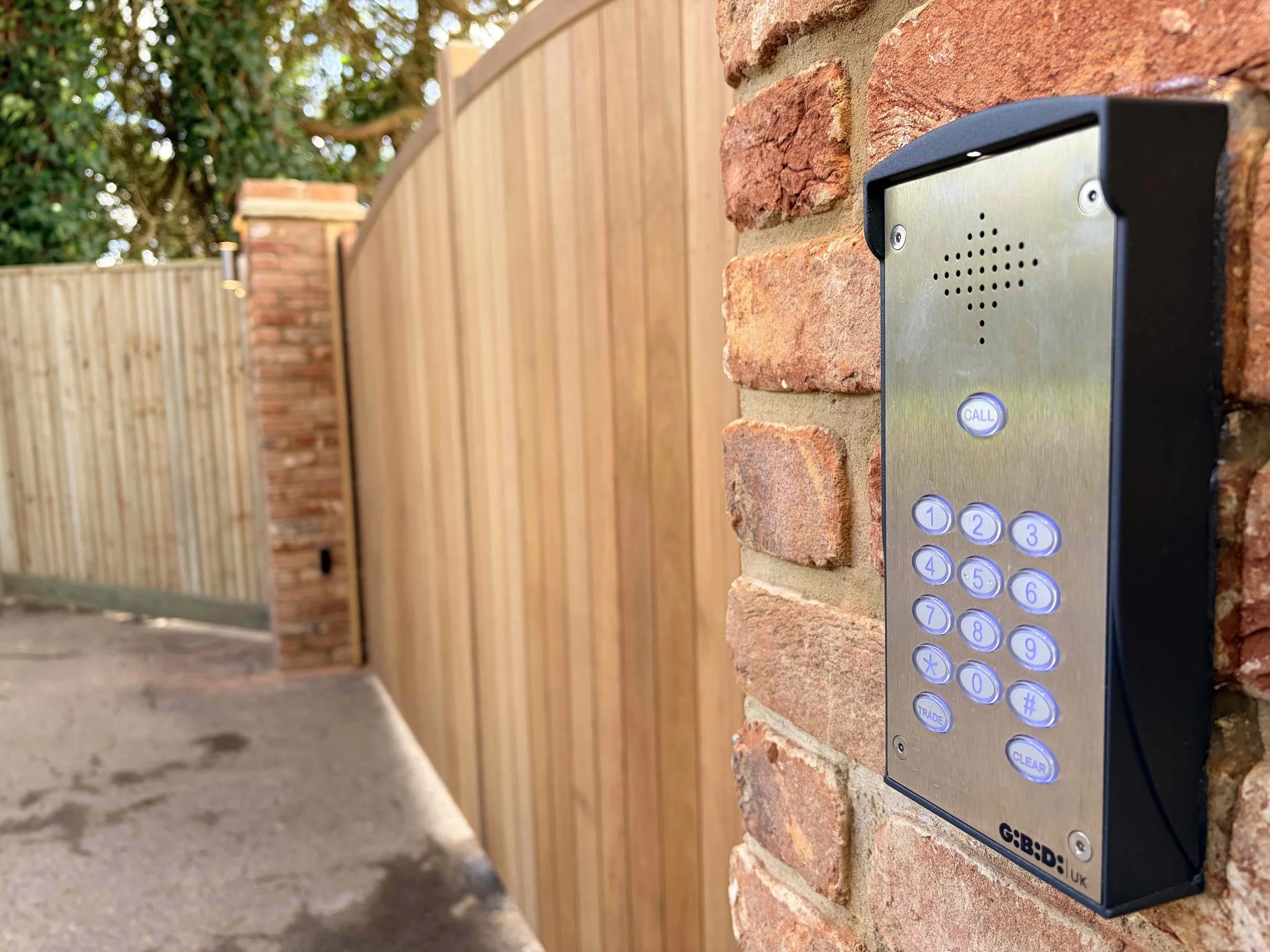 A close-up of a metallic intercom or access control keypad mounted on a brick wall, with a wooden fence and trees in the background.