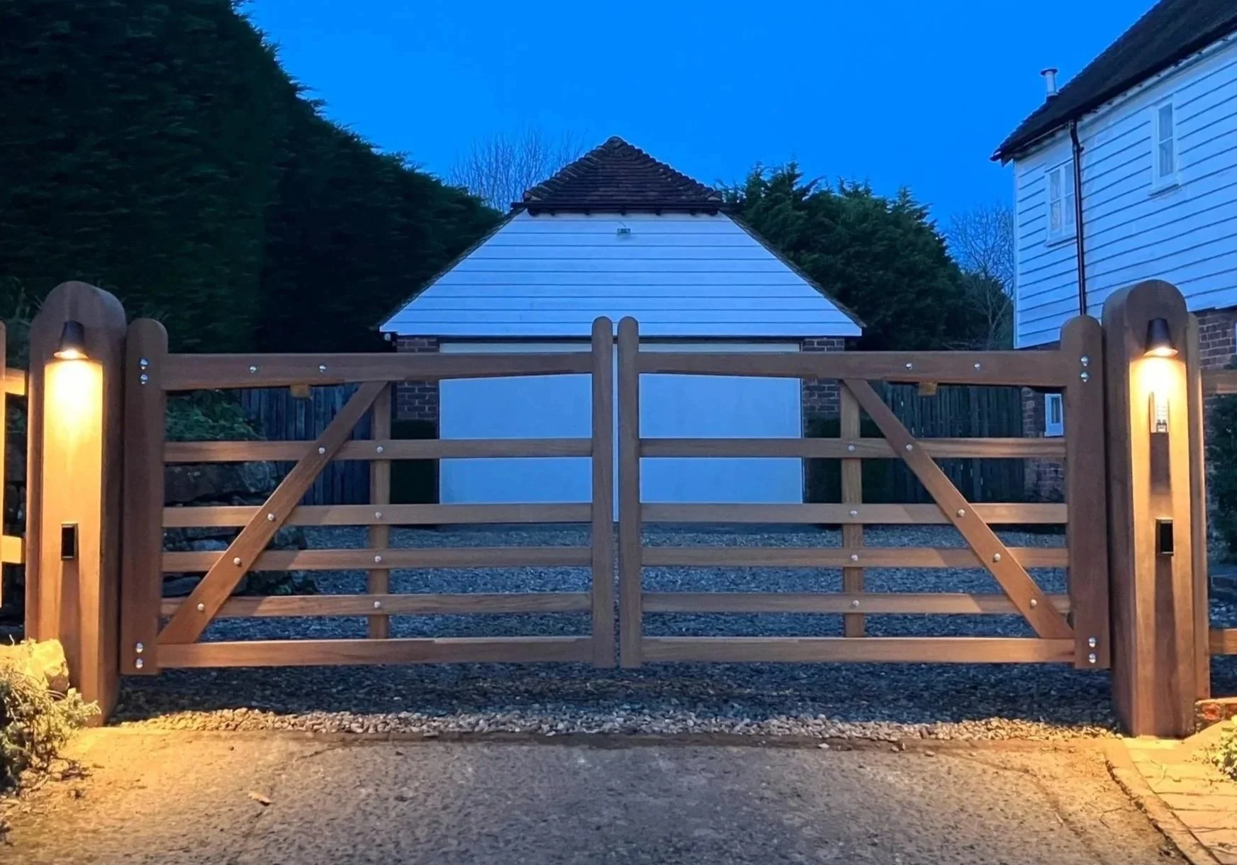 A wooden gate with two lamps on either side, leading to a backyard with a small building covered by a tiled roof, surrounded by trees and neighboring houses, during dusk.