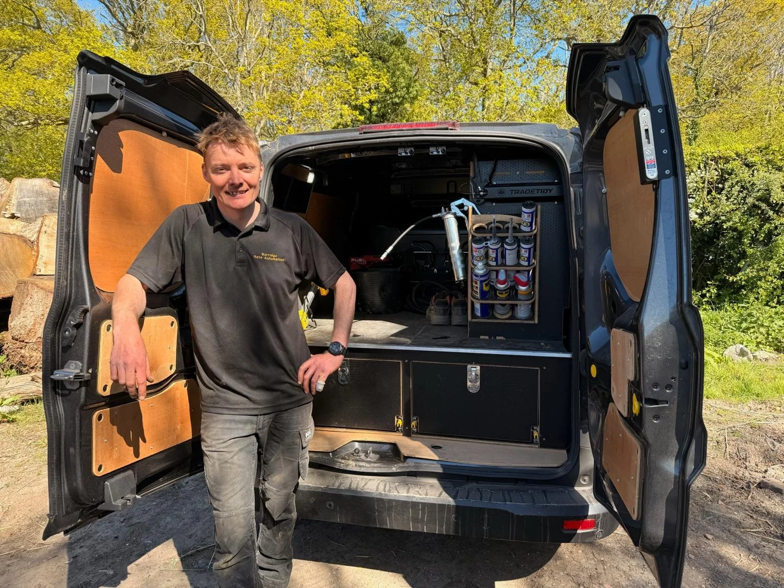 A man standing in front of an open work van with tools and supplies inside on a sunny day, with trees and a log pile in the background.